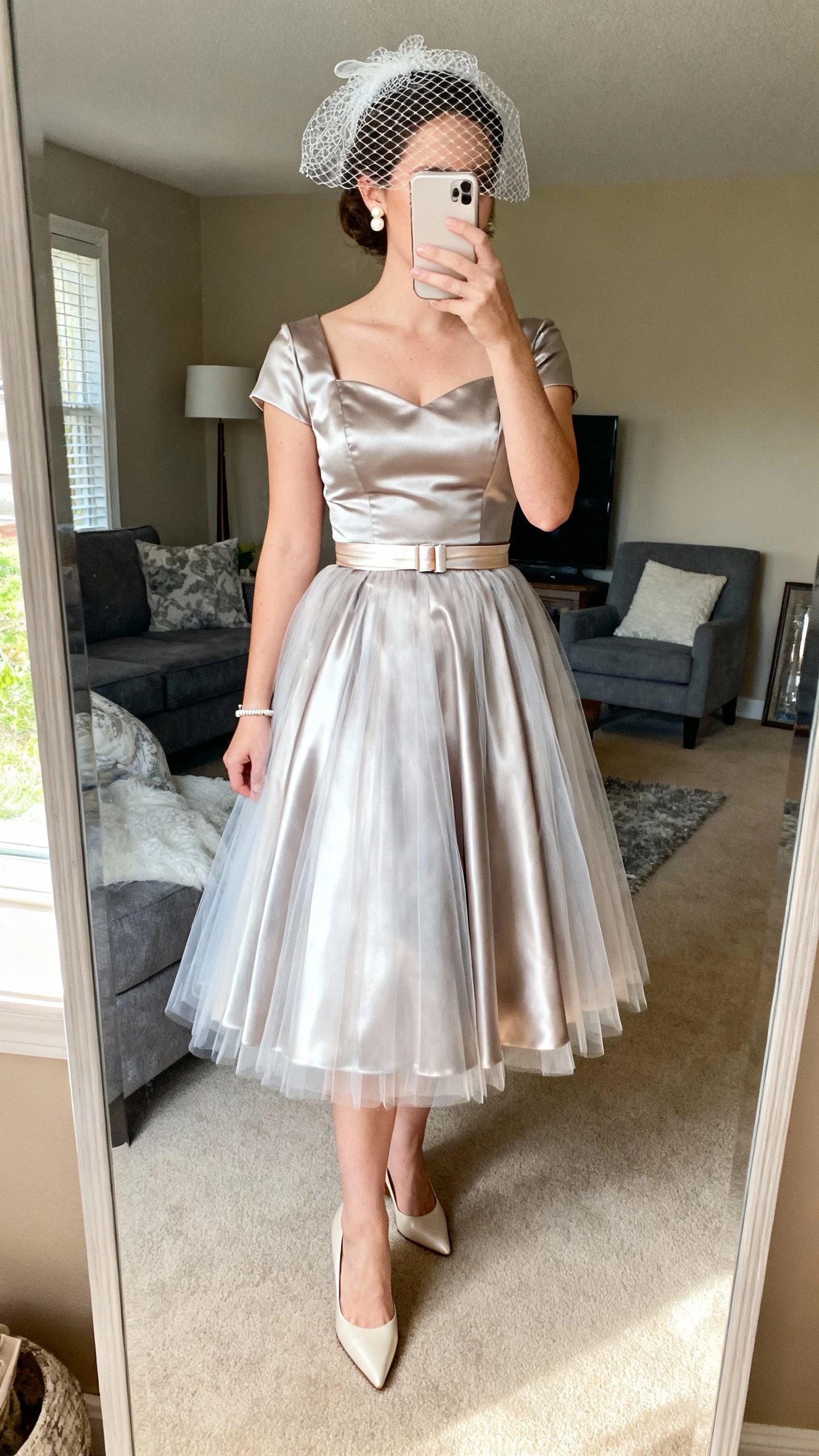 iPhone mirror selfie cropped at neck showing a woman in a 1950s tea-length satin dress with fitted bodice and tulle petticoat, pointed kitten heels, short birdcage veil, statement pearl studs, slim bracelet, ribbon belt at the waist, face not visible, living room full-length mirror, bright daylight, natural iPhone photo.