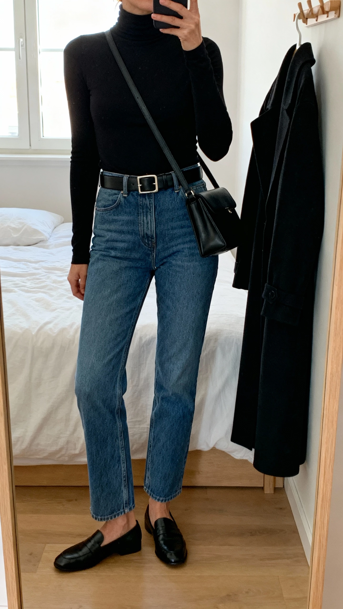 iPhone mirror selfie cropped at neck showing a woman in a black fitted mock neck, blue straight-leg jeans, black belt with subtle buckle, black loafers, black crossbody, optional black longline coat on hook; minimalist bedroom, face not visible, natural window light, natural iPhone photo quality.