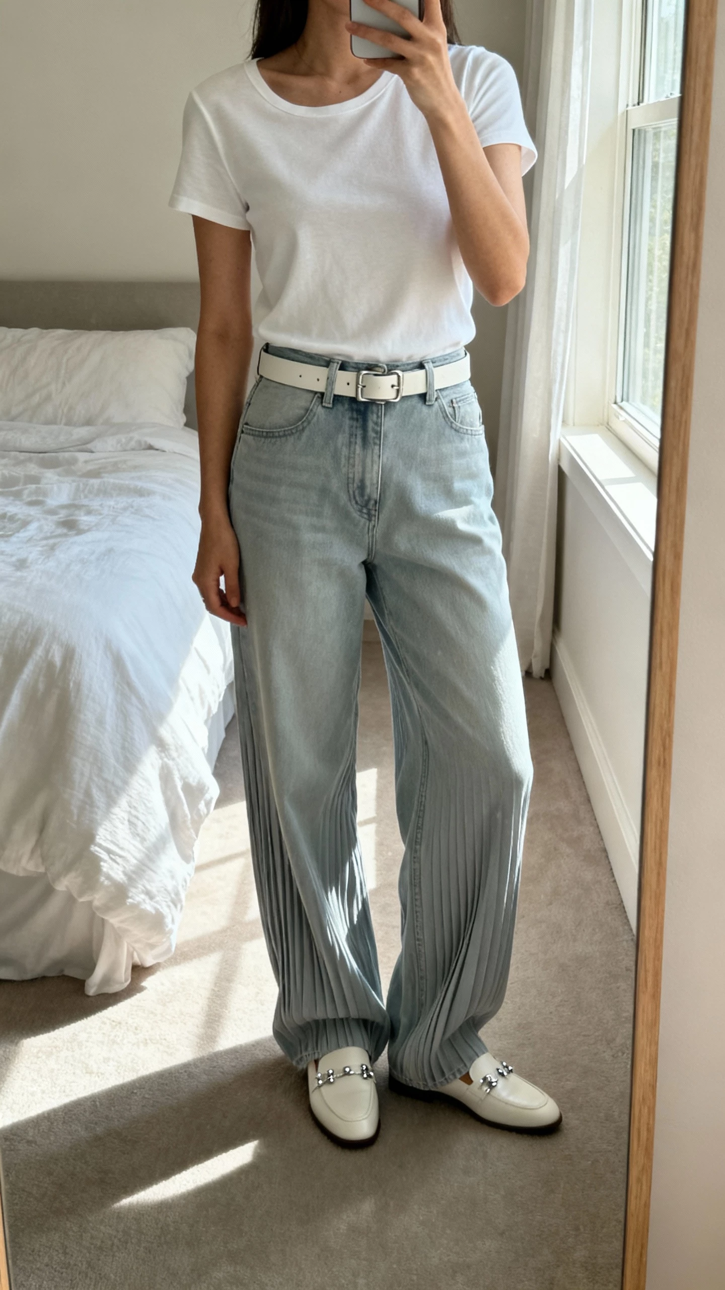 iPhone mirror selfie cropped at shoulders showing a woman in a minimalist monochrome outfit with a sleek tee, soft pleated baggy jeans, matching belt, clean loafers, subtle silver studs; bedroom mirror, face not visible, window light, natural iPhone photo.
