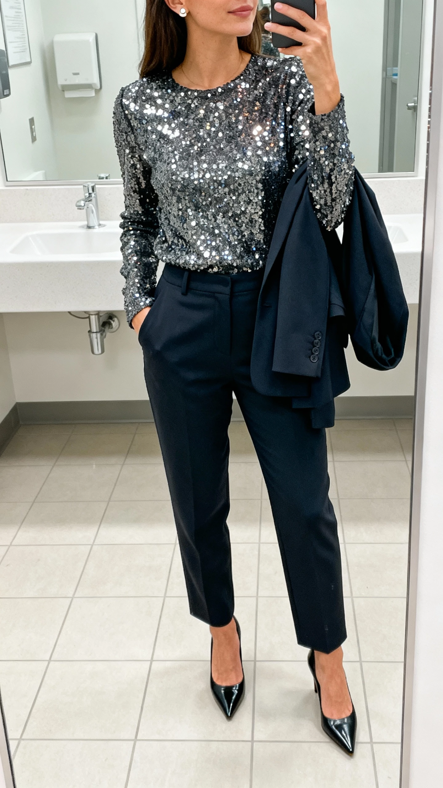 iPhone mirror selfie cropped at shoulders showing a woman wearing a sequined top tucked into tailored trousers, pointed heels, structured blazer draped over arm, and simple stud earrings, face not visible, office restroom mirror, bright indoor lighting, natural iPhone photo quality.