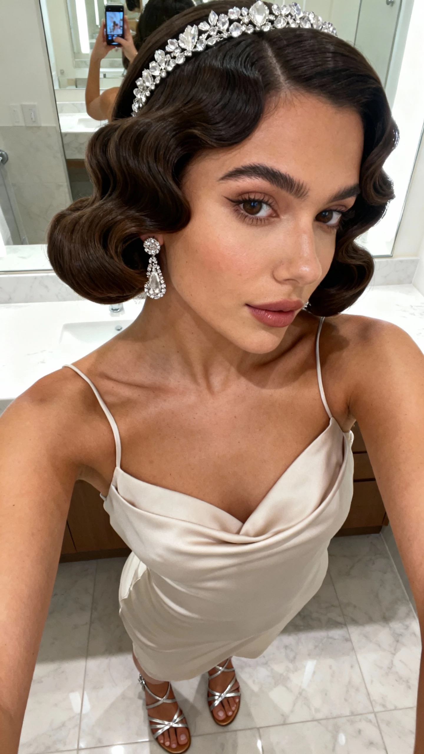 iPhone mirror selfie cropped at shoulders showing sleek Marcel waves with a crystal headband, bias-cut slip dress, rhinestone drop earrings, strappy metallic sandals on floor; face not visible, bathroom mirror, bright indoor lighting, casual iPhone photo.