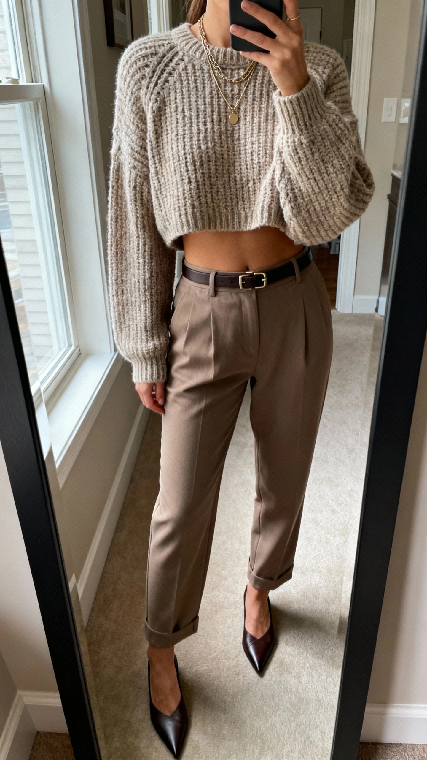 iPhone mirror selfie cropped at shoulders showing a woman in a cropped chunky knit with high-waisted trousers and pointed flats, layered necklaces and a slim belt, face not visible, apartment entryway mirror, morning window light, natural iPhone quality