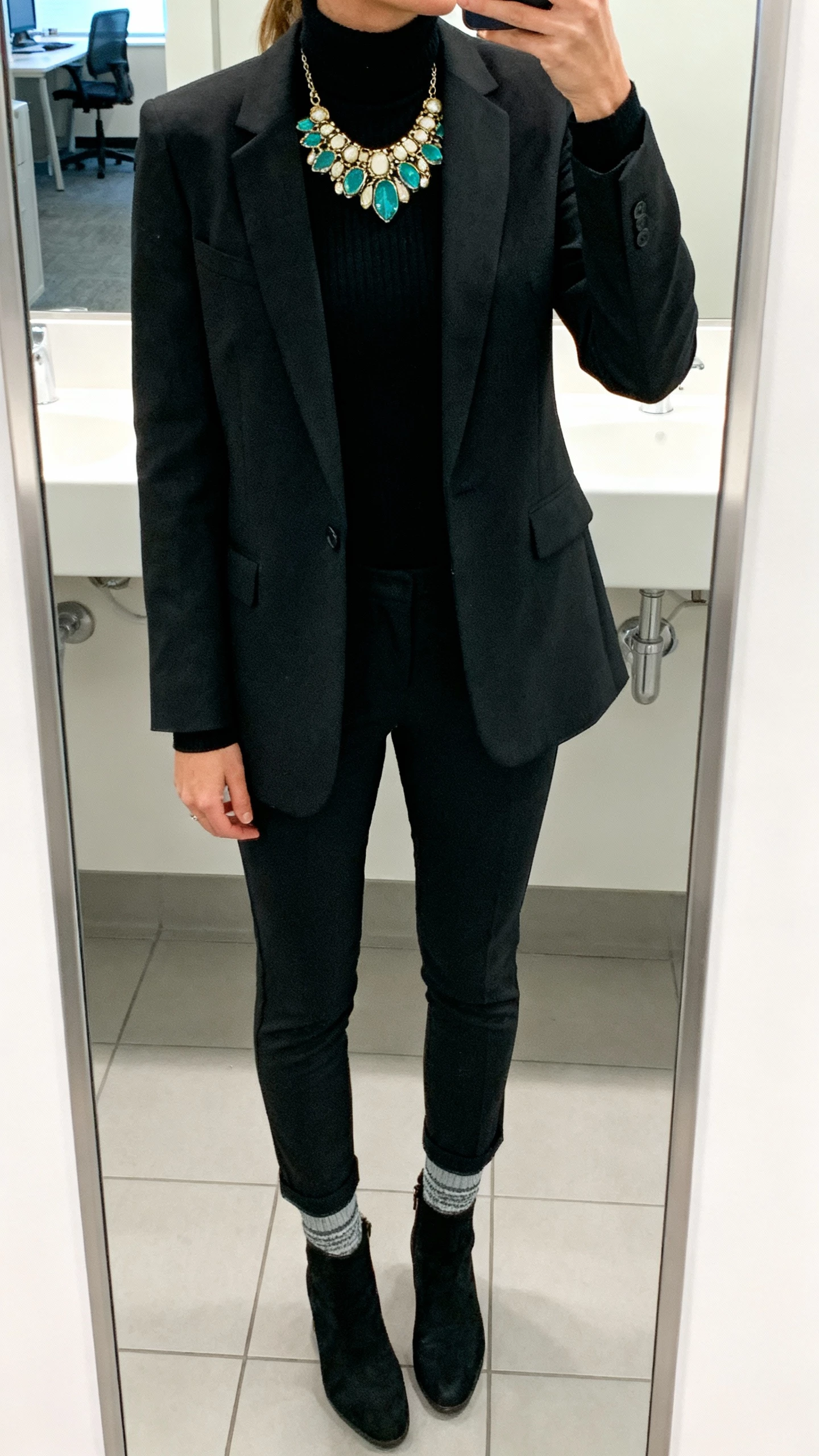 iPhone mirror selfie cropped at shoulders showing a woman wearing a bold statement necklace over a black turtleneck, tailored blazer, slim trousers, and sock boots, face not visible, office restroom mirror with bright indoor lighting, natural iPhone photo quality.