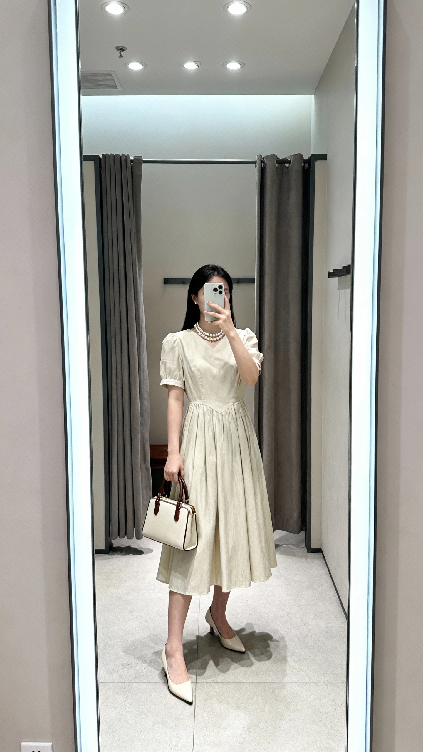 iPhone mirror selfie cropped at shoulders showing a woman in an A-line tea dress with vintage-inspired ease, kitten heels, pearl necklace, top-handle bag, face not visible, dressing room mirror, bright overhead lights, natural iPhone aesthetic.