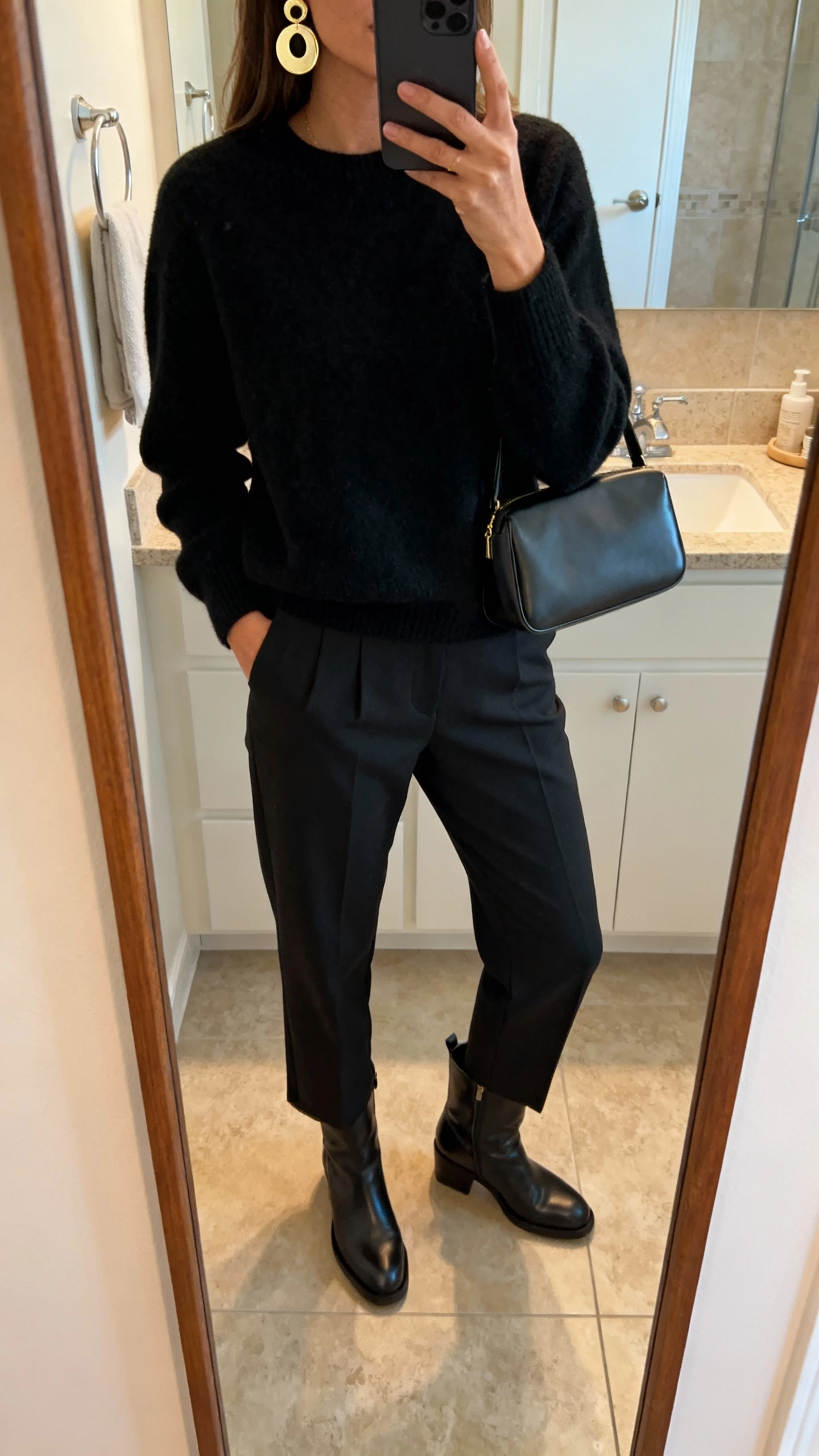 iPhone mirror selfie of a woman choosing one statement piece (bold earrings) with everything else minimal: simple black sweater, clean-cut trousers, classic boots, structured bag, face not visible, bathroom mirror, warm indoor light, iPhone photo quality.