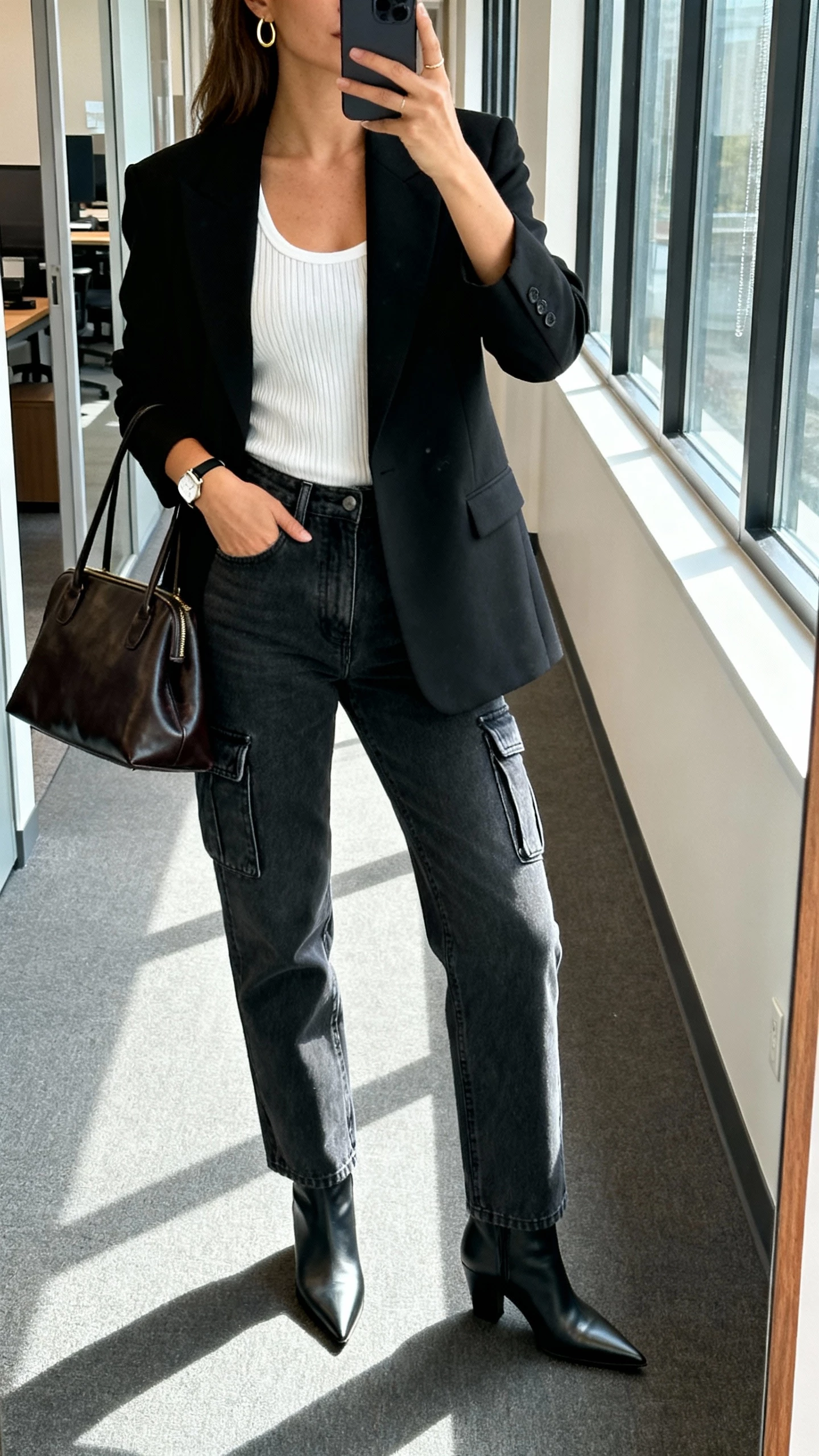iPhone mirror selfie of a woman in a black tailored blazer over a white ribbed tank, mid-rise straight cargo jeans, pointed-toe ankle boots, structured leather tote, minimal gold hoops and a slim watch, face not visible, cropped at neck, office hallway mirror, morning window light, natural iPhone photo quality.