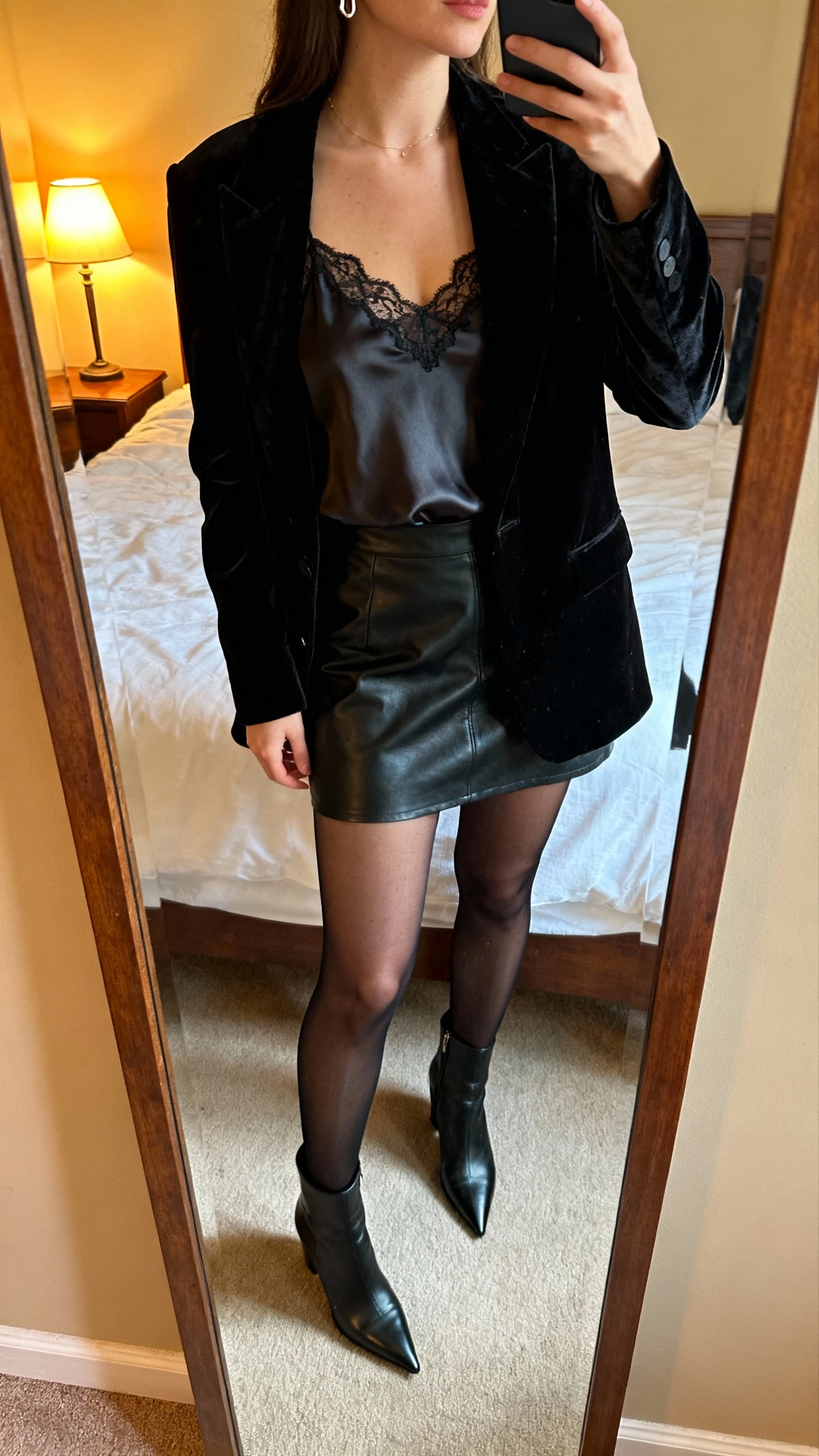 iPhone mirror selfie of a woman in a black velvet blazer over a silky lace camisole, mini skirt in leather, sheer black tights, pointed-toe heeled ankle boots, delicate earrings peeking, face not visible, cropped at neck, bedroom mirror with warm lamp light, natural iPhone photo quality.