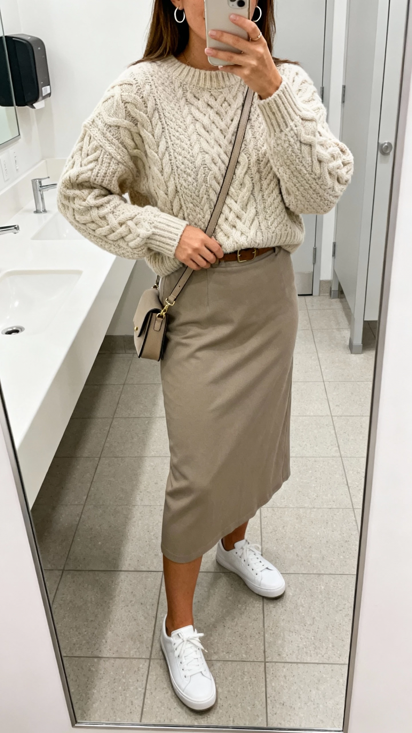 iPhone mirror selfie of a woman in a cable-knit sweater tucked into a midi skirt, white low-top sneakers, crossbody bag, subtle hoops, face not visible, office restroom mirror, bright indoor light, natural iPhone photo quality