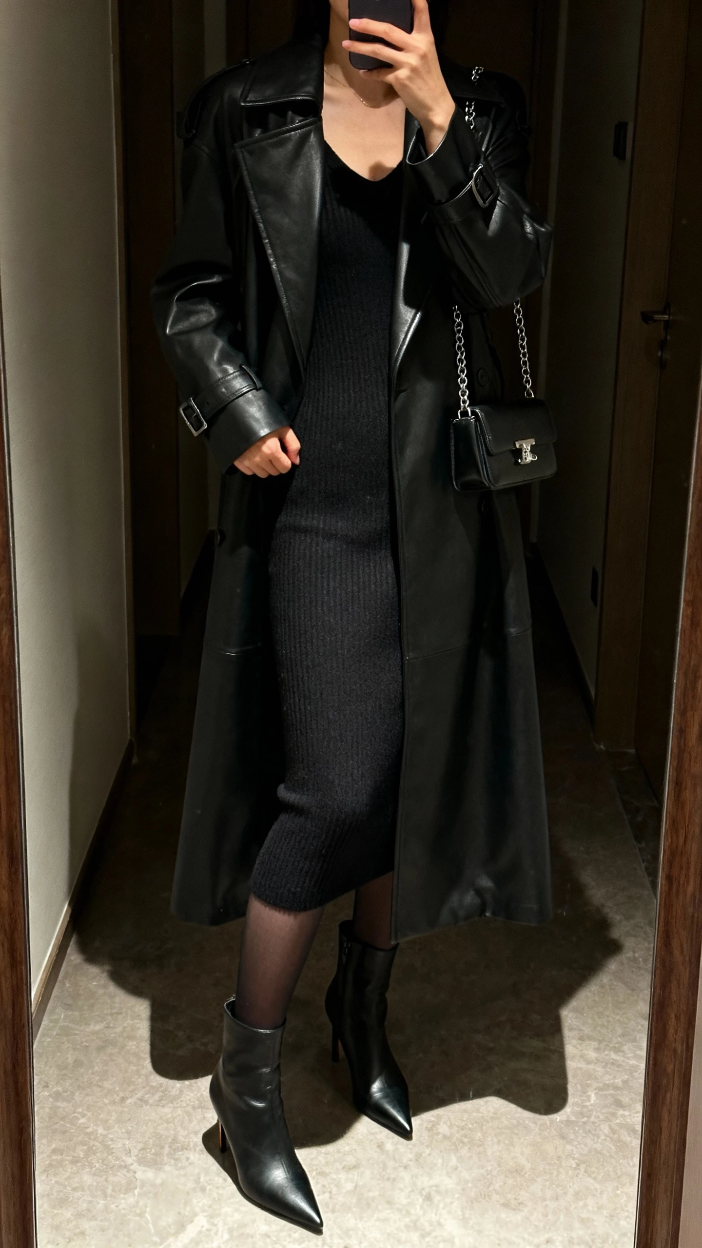 iPhone mirror selfie of a woman in a calf-length black leather trench over a body-skimming black knit dress, opaque tights, pointed heeled boots, compact shoulder bag with chain strap; dim hallway mirror, cropped at neck, face not visible, evening indoor lighting, natural iPhone photo quality.