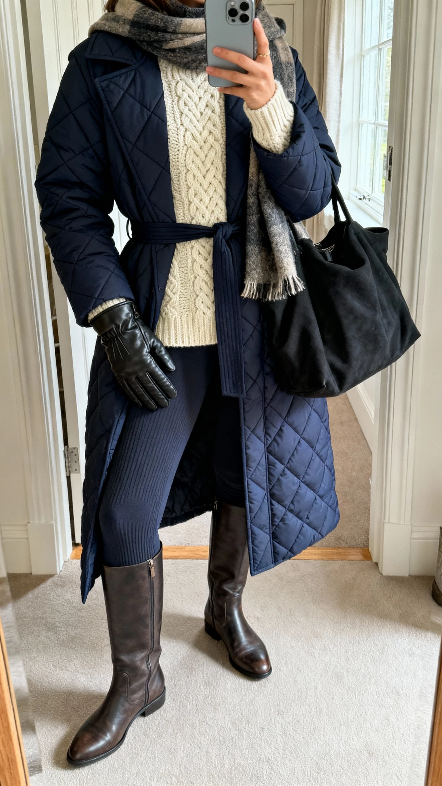 iPhone mirror selfie of a woman in a calf-length navy quilted coat (belted) over a chunky cream fisherman sweater, thick thermal leggings, knee-high flat leather boots, slouchy black tote, wool blanket scarf and leather tech gloves tucked in; entryway mirror, cropped at shoulders, face not visible, morning window light, iPhone photo quality.