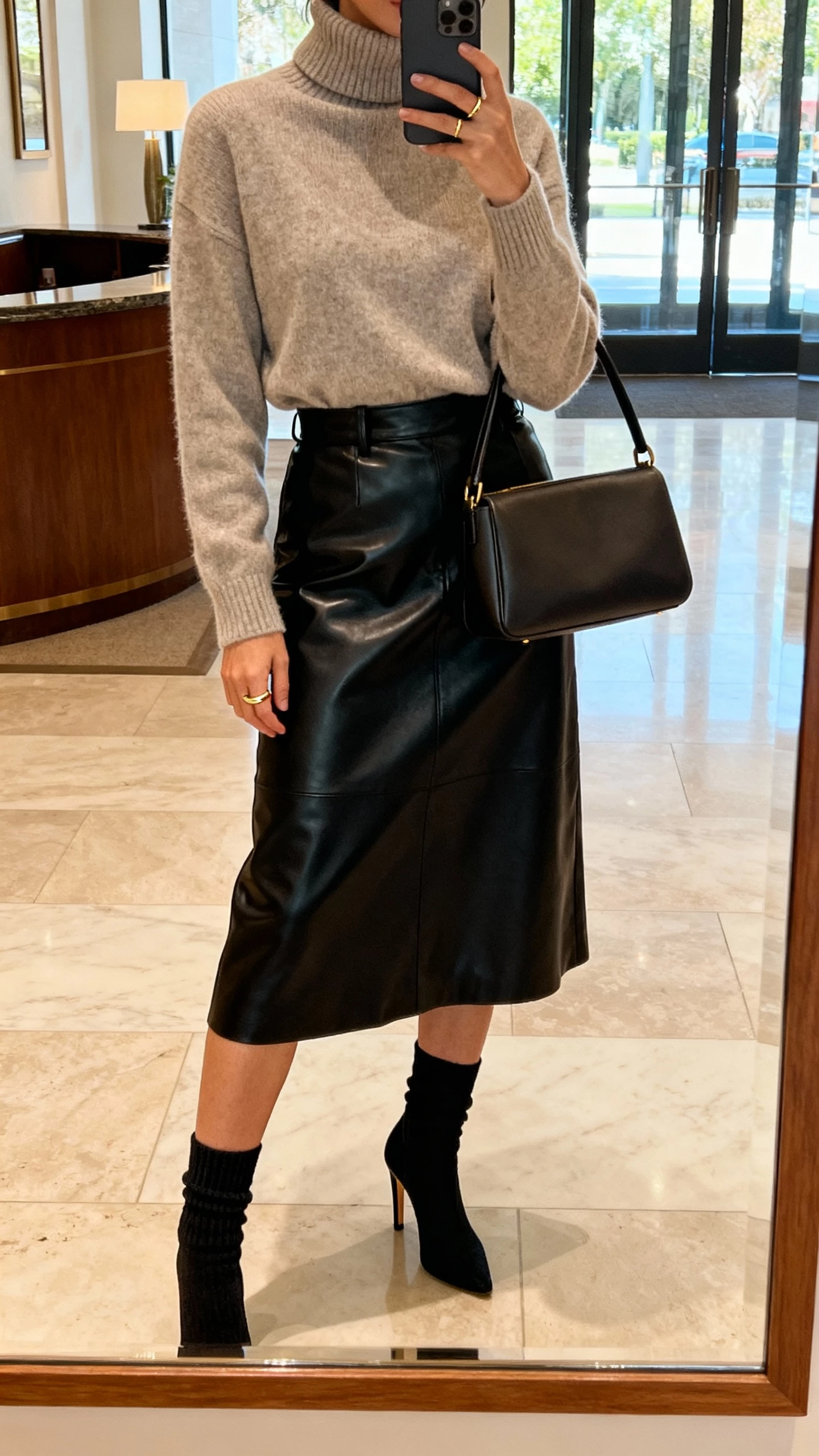 iPhone mirror selfie of a woman in a cashmere turtleneck tucked into a black leather midi skirt, heeled sock boots, minimal gold ring, structured top-handle bag, face not visible, lobby mirror, afternoon natural light, iPhone photo quality.