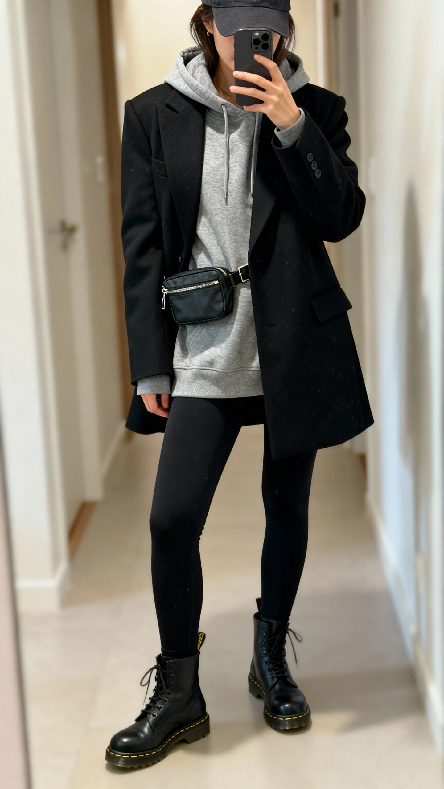 iPhone mirror selfie of a woman in a charcoal hoodie under a tailored black coat, black leggings, black Doc Martens, baseball cap, and a small belt bag, face not visible, cropped at shoulders, hallway mirror with soft indoor lighting, natural iPhone photo quality.