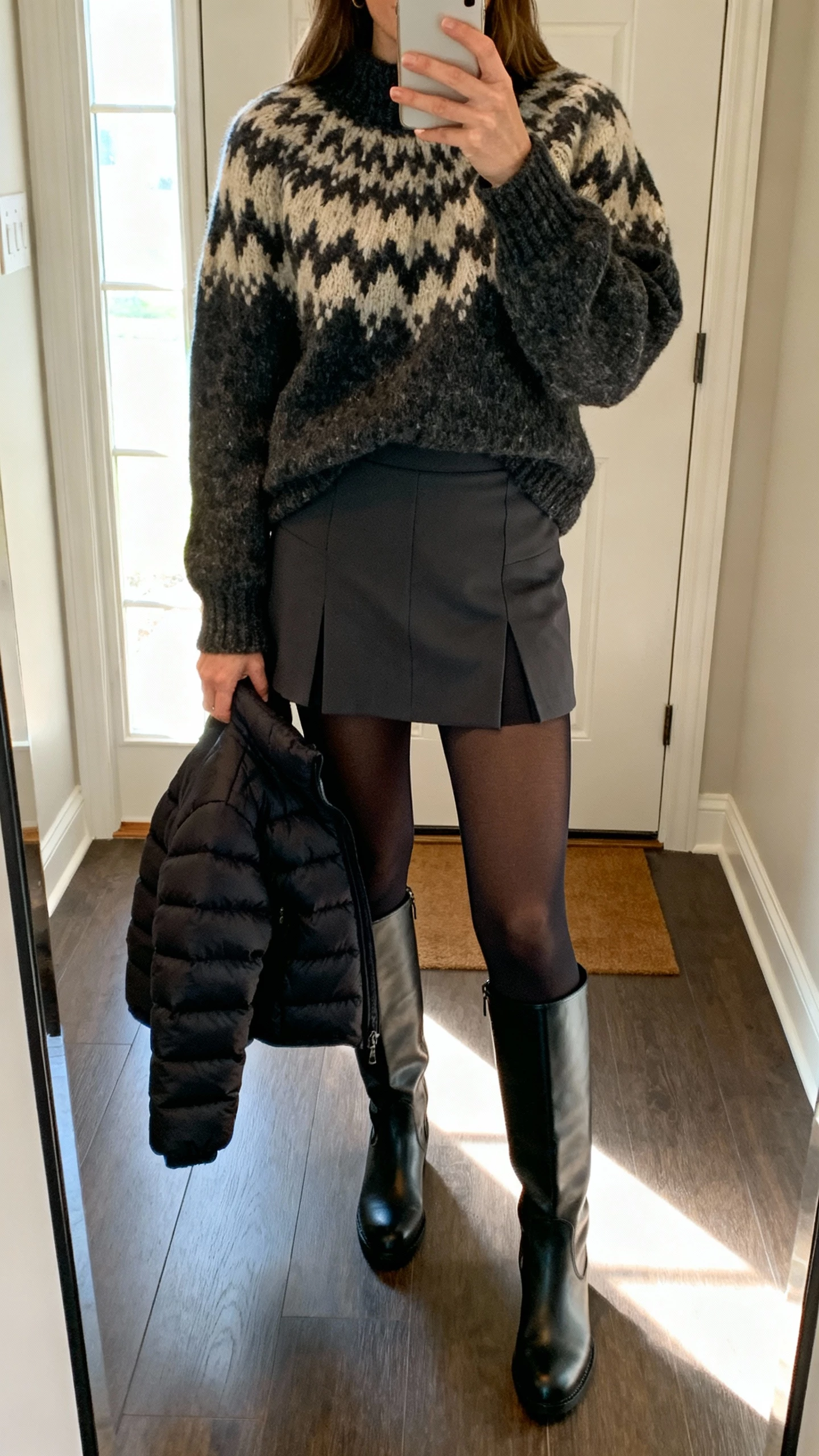 iPhone mirror selfie of a woman in a chunky statement sweater, structured mini skort, thermal tights, over-the-knee boots, short puffer jacket in hand, face not visible, cropped at neck, entryway mirror with daylight from door, natural iPhone photo quality.