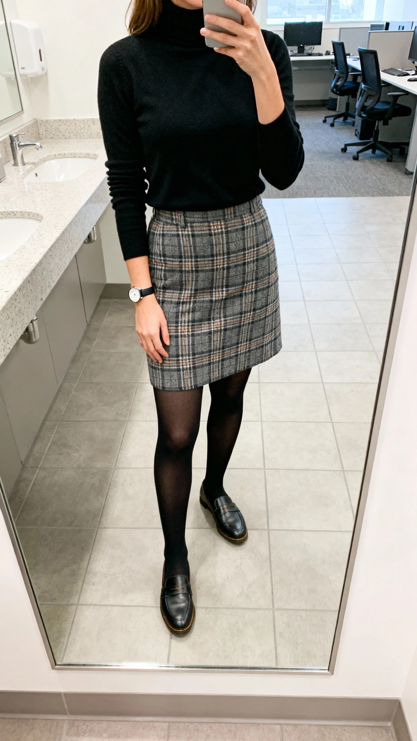 iPhone mirror selfie of a woman in a classic black turtleneck, plaid skirt, loafers, and black tights with a simple watch, face not visible, office restroom mirror, bright indoor lighting, natural iPhone look.
