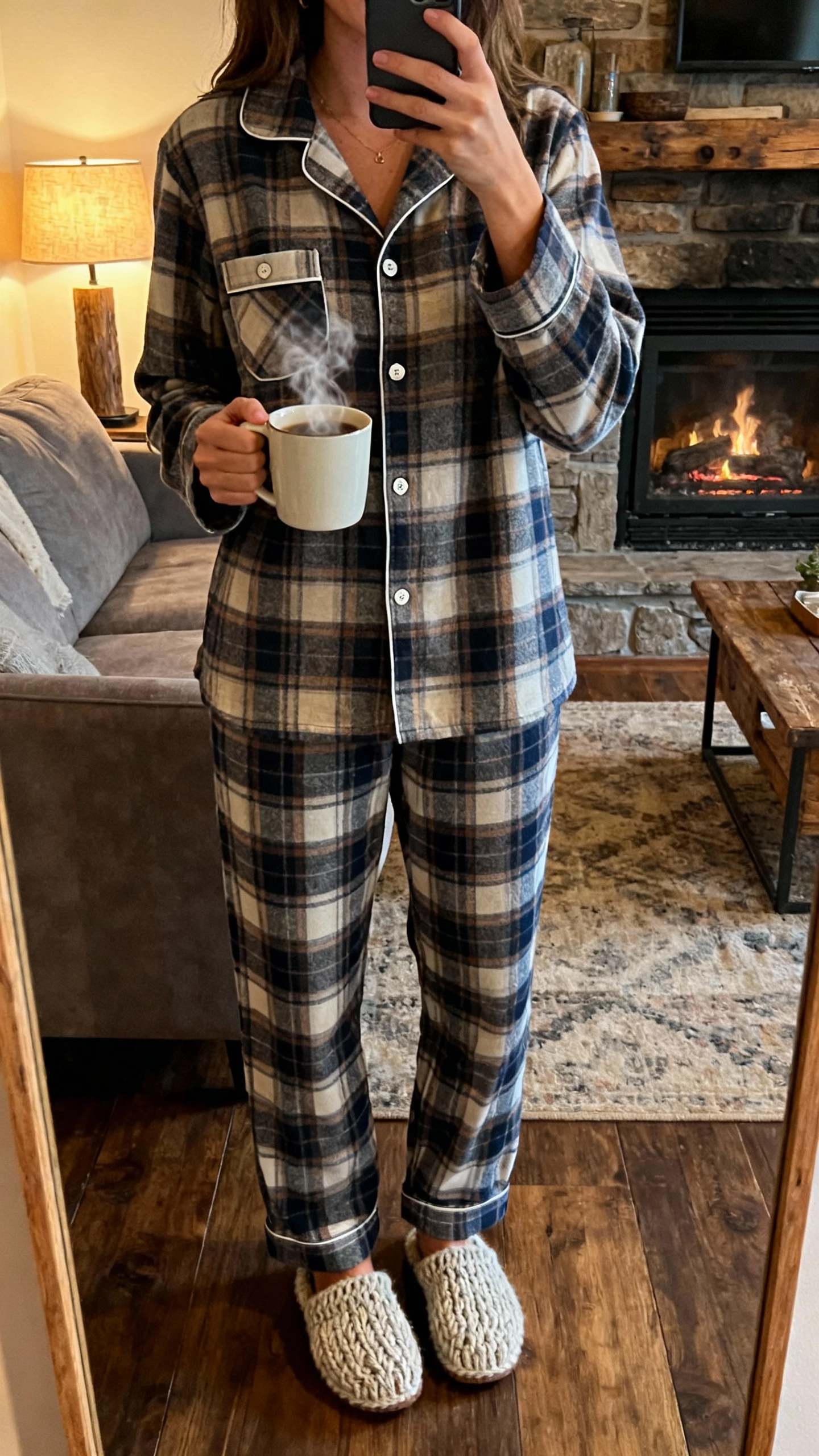 iPhone mirror selfie of a woman in a classic flannel pajama set (button-up shirt with chest pocket and matching plaid pants) plus knit slippers and a steaming mug, face not visible, rustic living room near a fireplace, soft evening lamp light, natural iPhone photo.