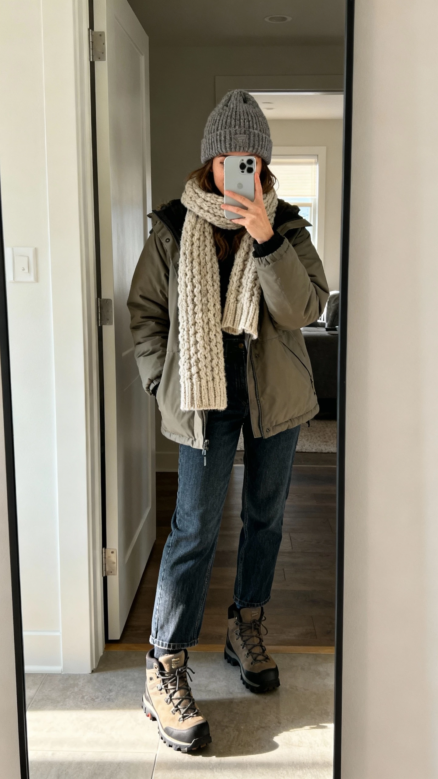 iPhone mirror selfie of a woman in a cozy parka with a chunky scarf, straight-leg denim, hiking-style boots, knit beanie, face not visible, entryway mirror with soft morning light, natural iPhone photo.