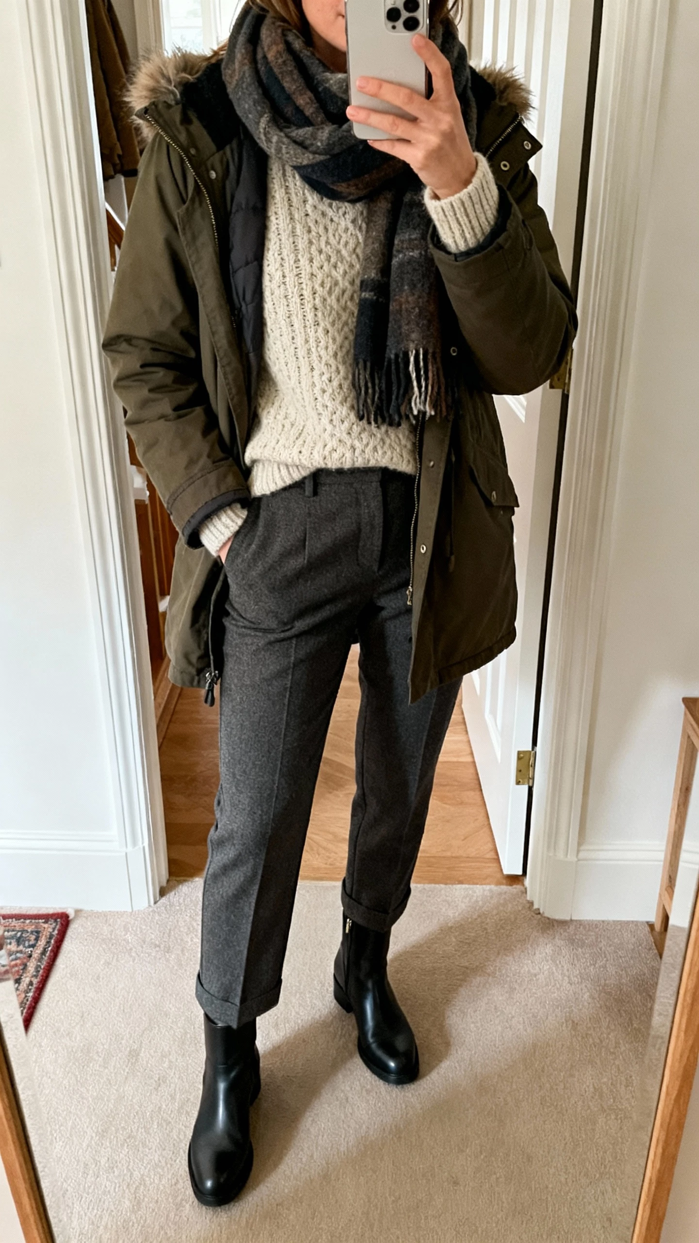 iPhone mirror selfie of a woman in a cozy, ready-for-winter look summarizing the vibe: layered knit, tailored bottoms, practical outerwear, scarf, and boots, face not visible, entryway mirror, soft natural light, natural iPhone photo quality.