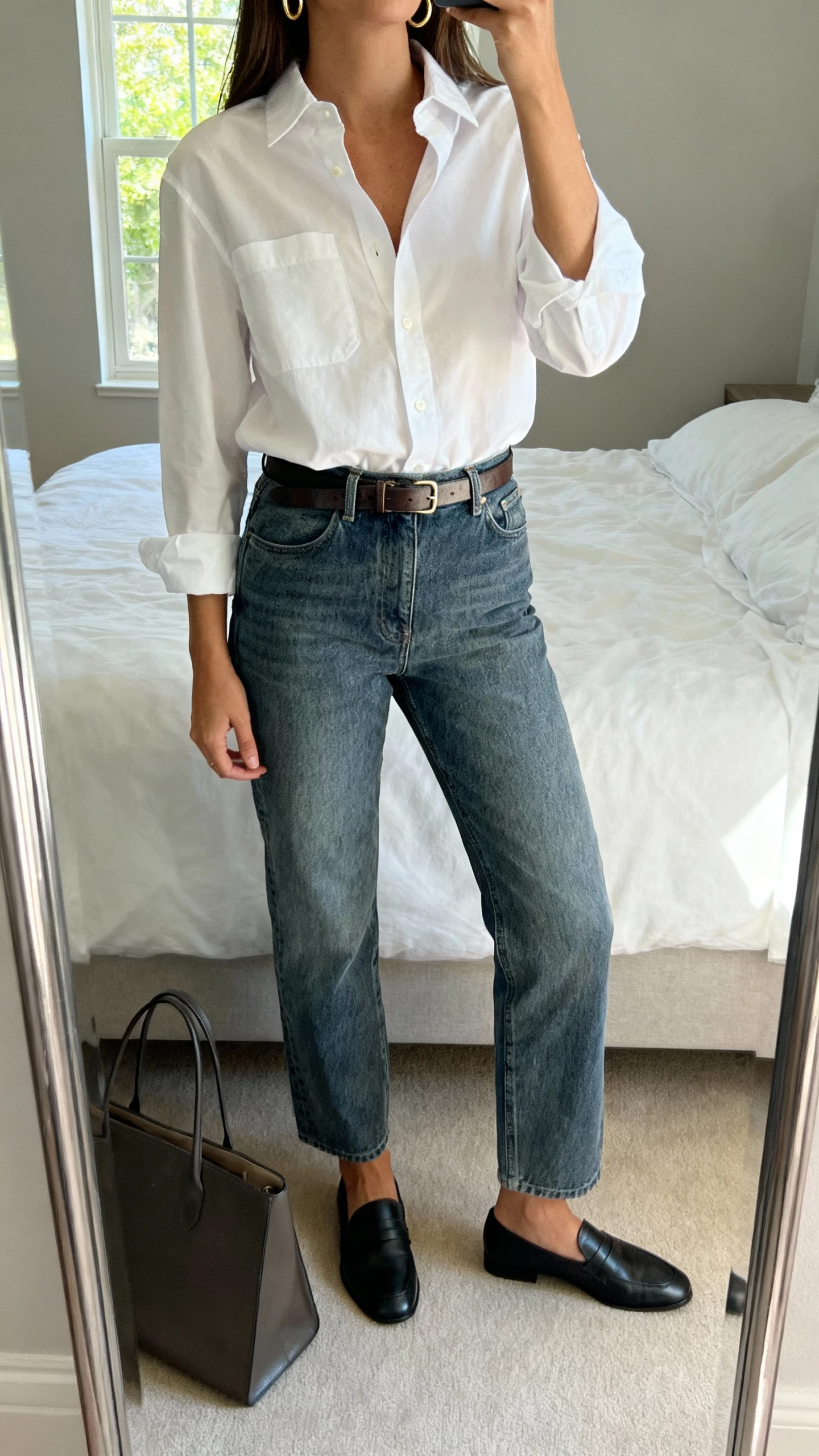 iPhone mirror selfie of a woman in a crisp white button-down tucked into mid-wash straight-leg jeans, slim leather belt, minimal gold hoops, black loafers, structured tote on floor, face not visible, cropped at neck, bedroom full-length mirror, natural window light, natural iPhone photo quality