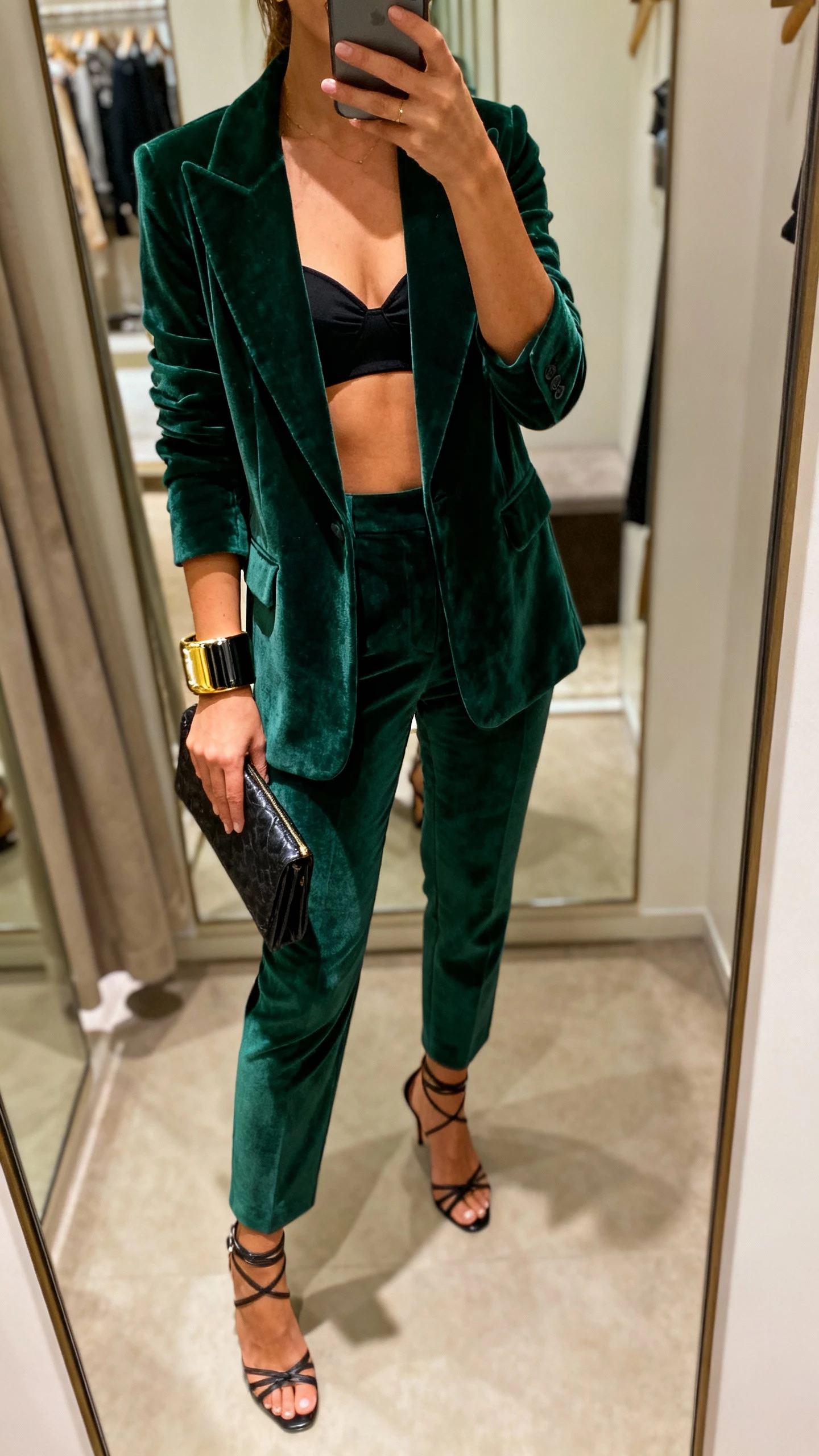 iPhone mirror selfie of a woman in a deep green velvet suit (blazer + tailored trousers) with a black bustier top, strappy heels, bold cuff bracelet, clutch, face not visible, boutique fitting-room mirror, warm indoor lighting, natural iPhone aesthetic.