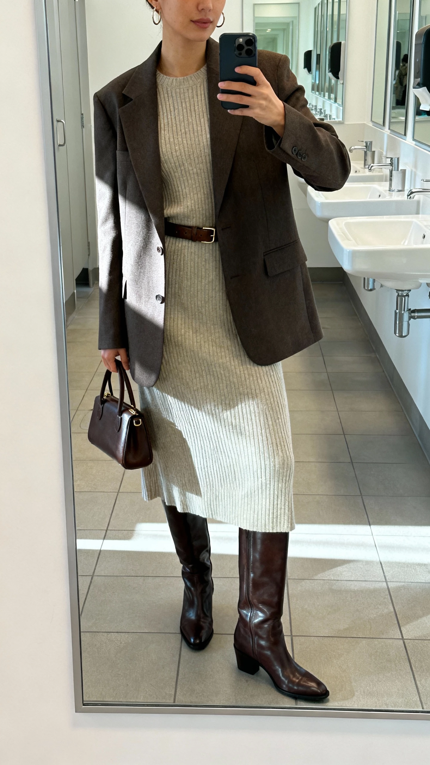 iPhone mirror selfie of a woman in a downtown tailored look: structured blazer over a ribbed knit midi dress, slick knee-high leather boots, slim leather belt, small top-handle bag, subtle hoop earrings, face not visible, office restroom full-length mirror, bright morning light, natural iPhone photo quality