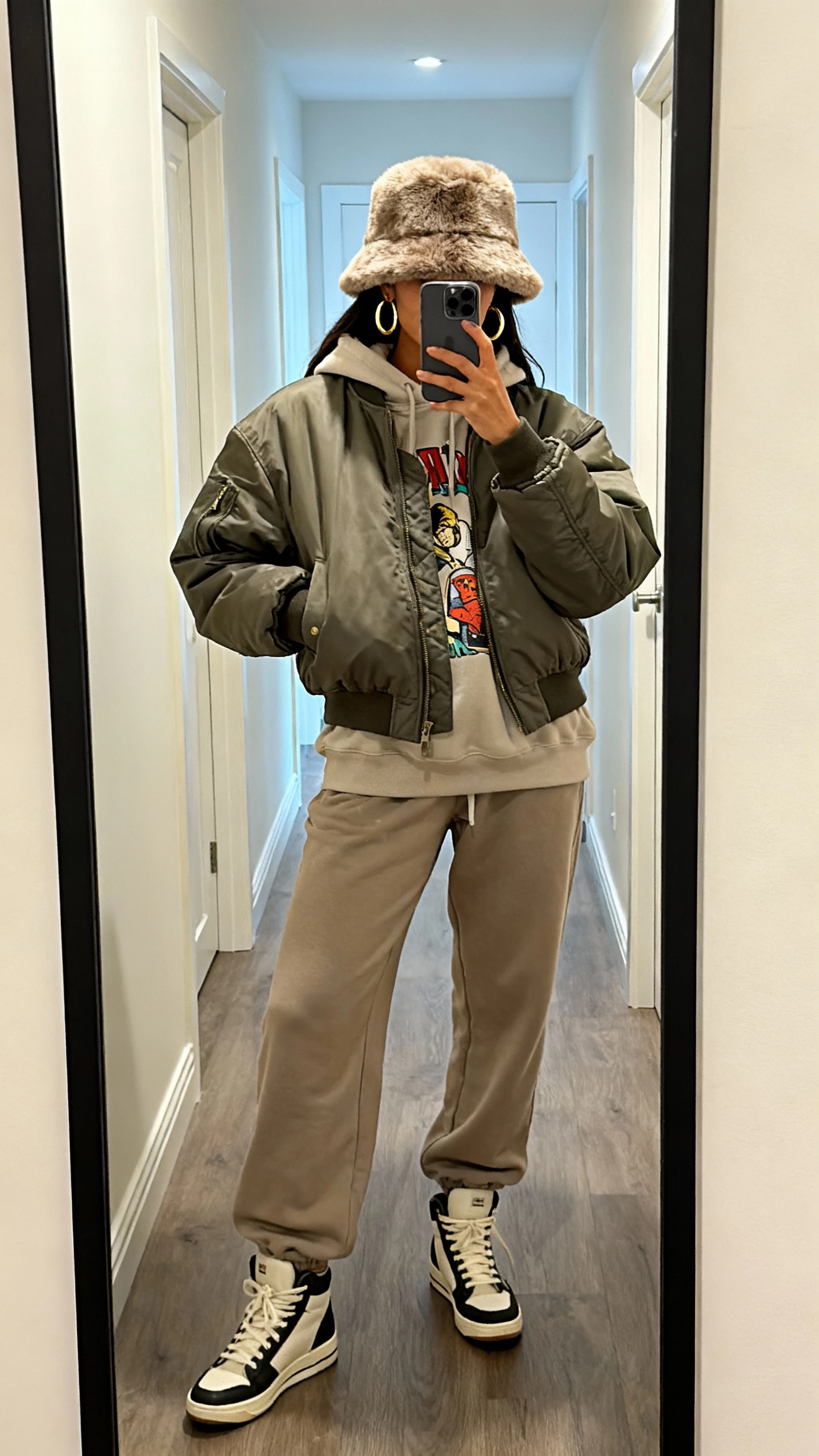 iPhone mirror selfie of a woman in a faux-fur bucket hat, oversized bomber, graphic hoodie, relaxed joggers, high-top sneakers, and bold hoop earrings; face not visible, hallway mirror, soft indoor lighting, natural iPhone photo quality.