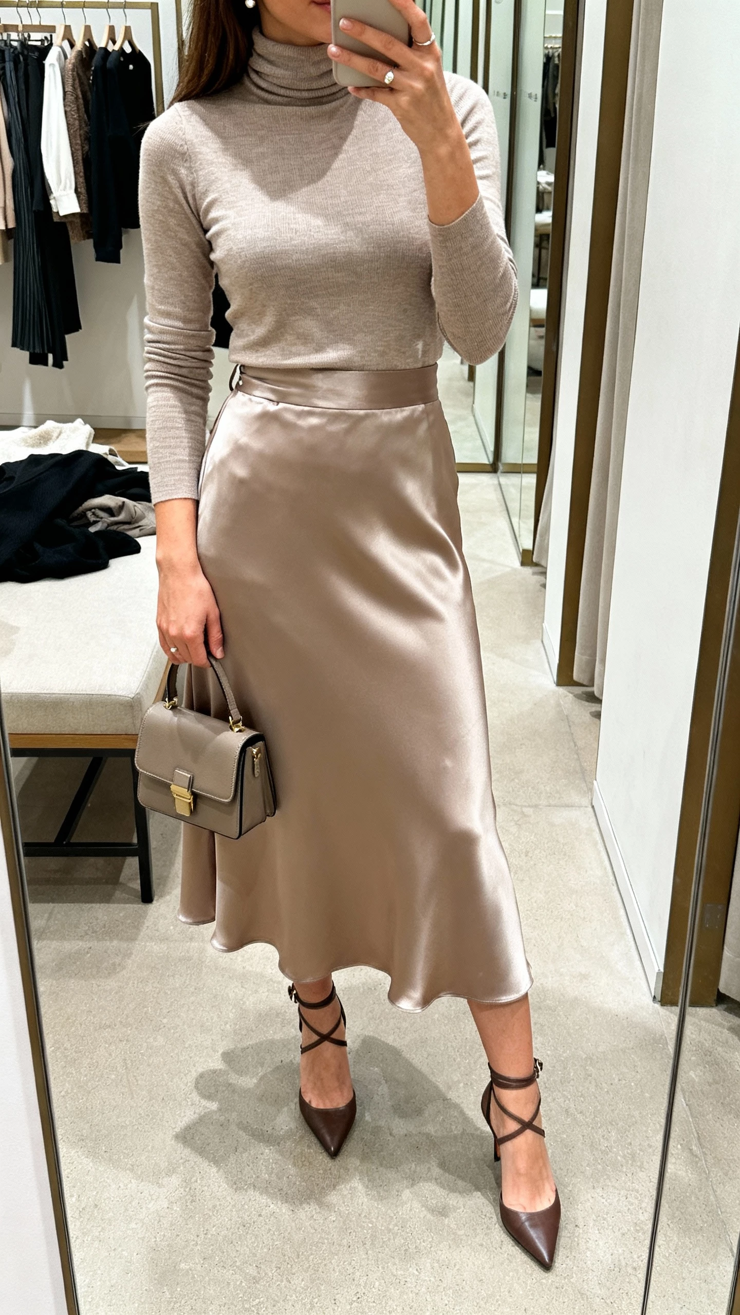 iPhone mirror selfie of a woman in a fitted turtleneck tucked into a satin midi skirt, ankle strap heels, structured mini bag, simple studs, face not visible, cropped at shoulders, boutique dressing room mirror, soft indoor lighting, natural iPhone photo quality