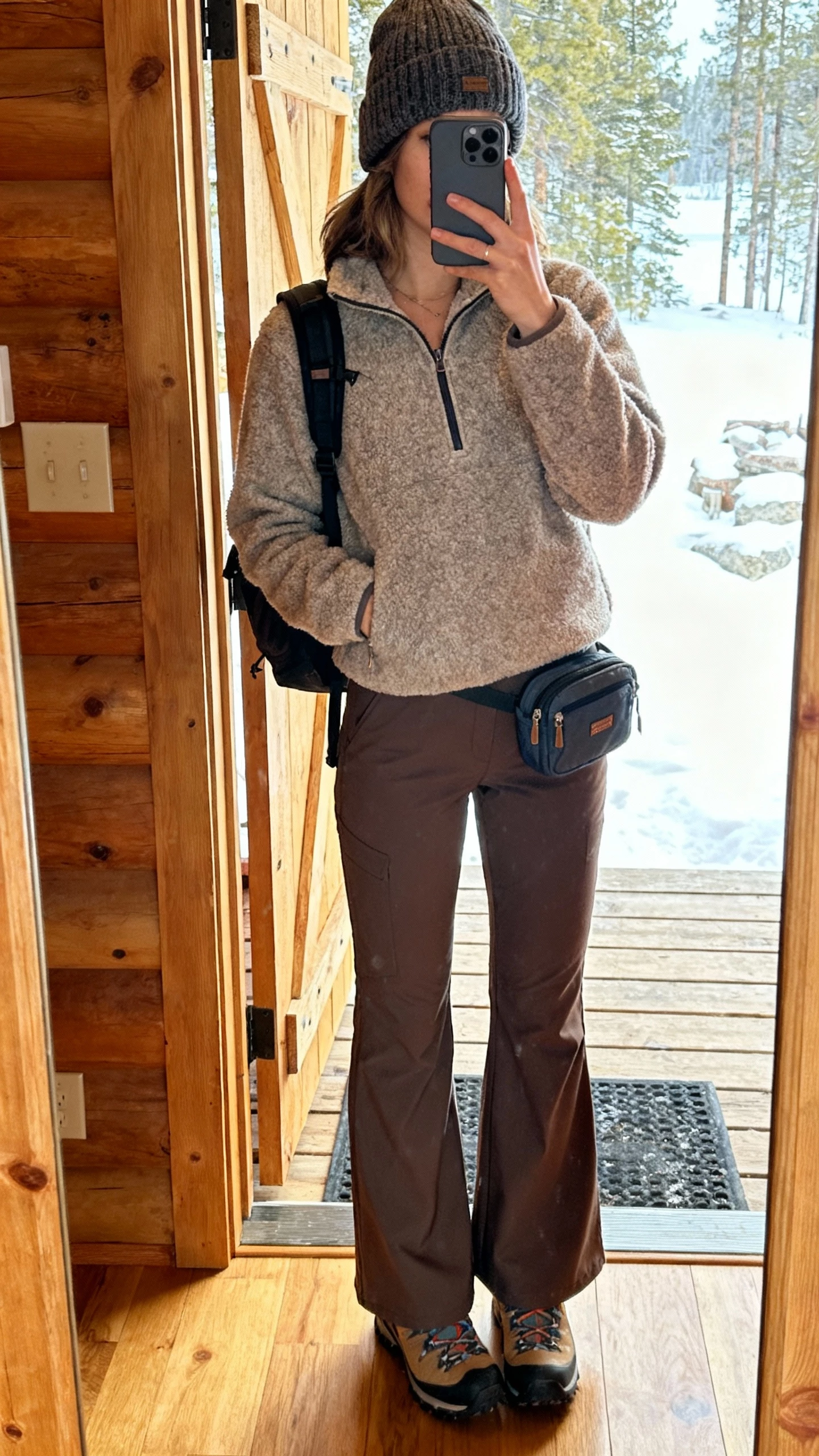 iPhone mirror selfie of a woman in a fleece half-zip, high-rise flare pants, hiking boots, wool beanie, mini backpack, face not visible, cabin entryway mirror, bright natural light, natural iPhone photo quality