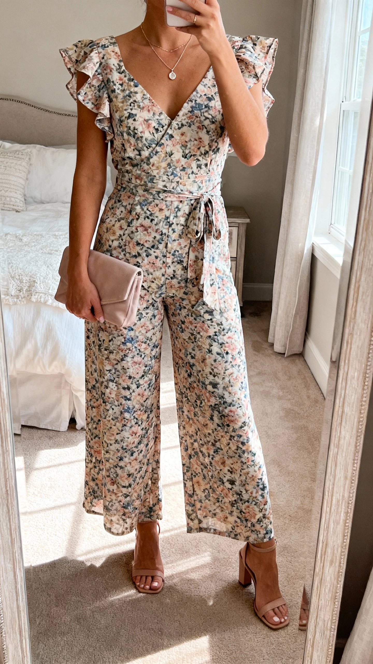iPhone mirror selfie of a woman in a floral wide-leg jumpsuit with flutter sleeves, dainty pendant necklace, soft clutch, heeled sandals, face not visible, bedroom full-length mirror with morning window light, natural iPhone photo quality