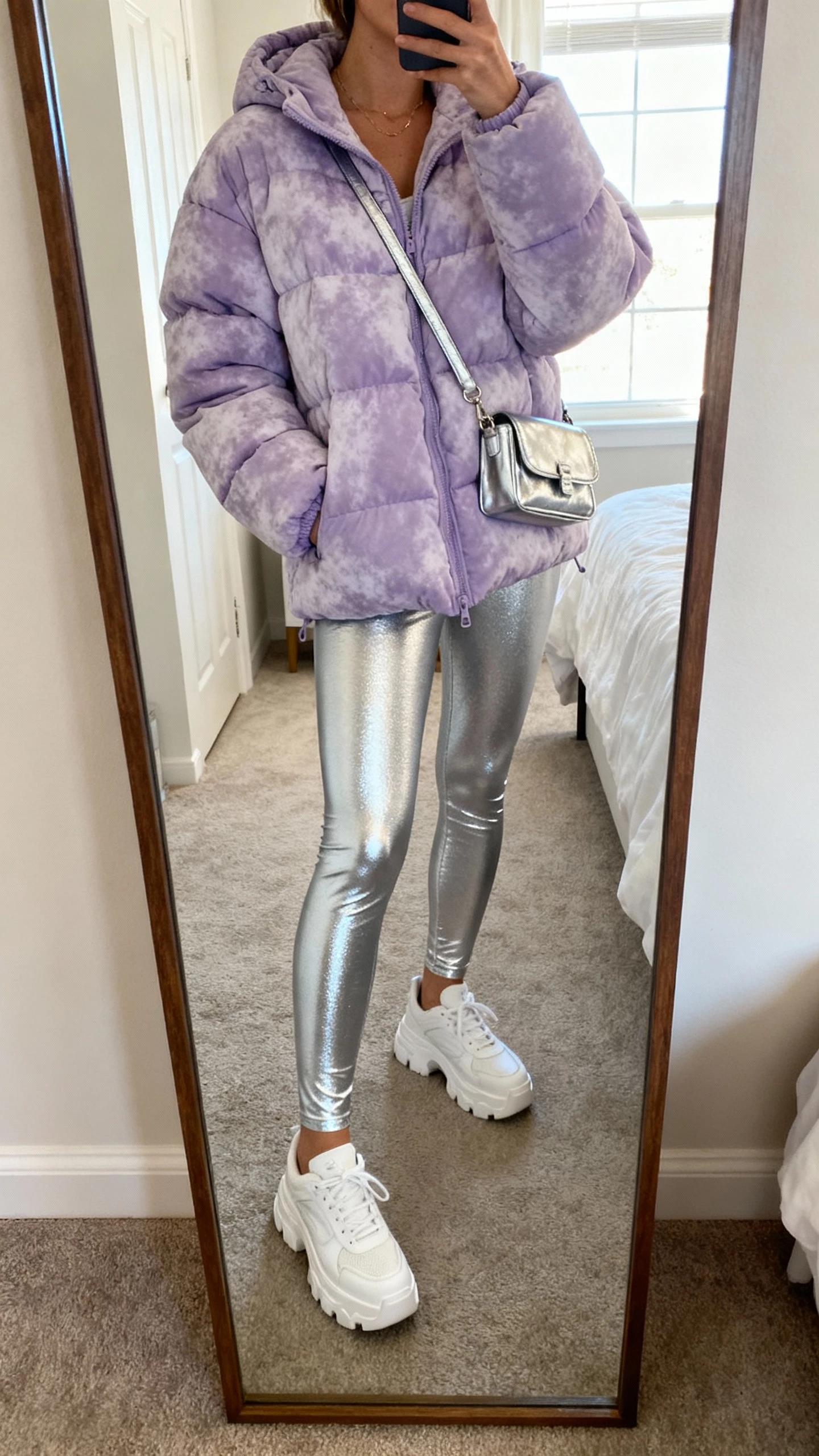 iPhone mirror selfie of a woman in a frosted lilac puffer jacket, liquid-silver leggings, white chunky sneakers, and a small metallic crossbody, face not visible, cropped at neck, bedroom full-length mirror with morning window light, natural iPhone photo quality.