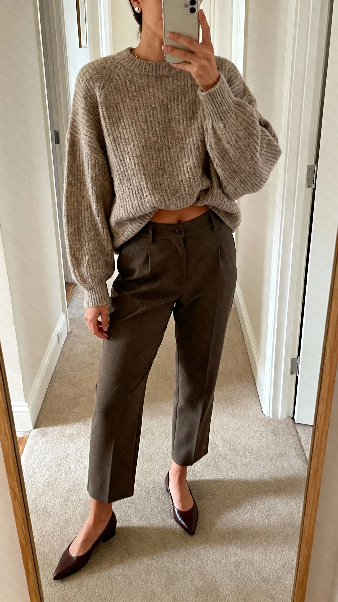 iPhone mirror selfie of a woman in a half-tucked oversized knit sweater with tailored trousers and pointed loafers, simple studs, face not visible, cropped at neck, hallway mirror, soft morning light, natural iPhone photo.