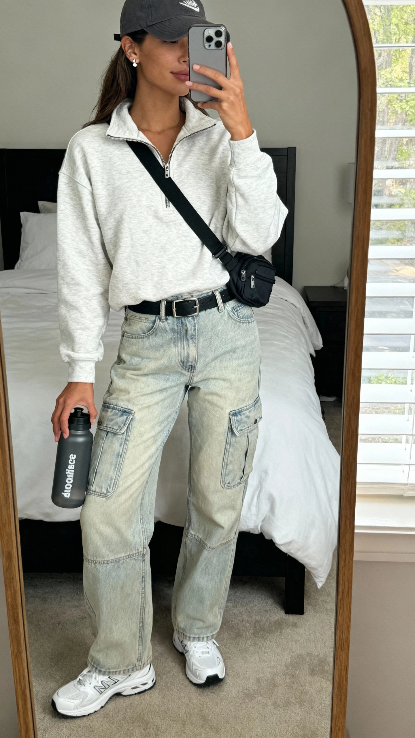iPhone mirror selfie of a woman in a half-zip sweatshirt, relaxed light-wash cargo jeans, clean white sneakers, baseball cap, crossbody belt bag, subtle stud earrings, and a gym-to-street water bottle in hand, face not visible, bedroom mirror, soft natural window light, casual iPhone photo.