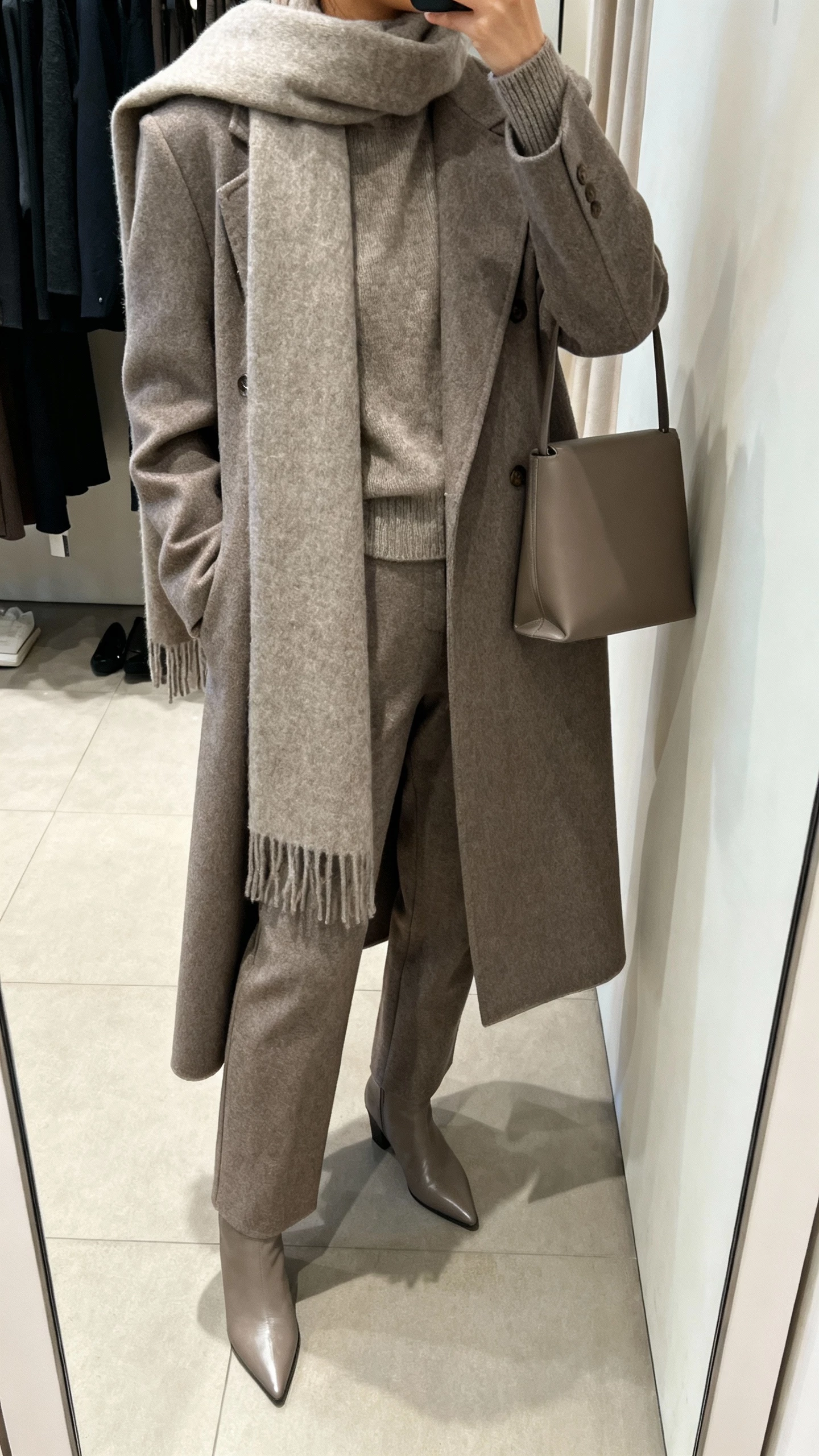 iPhone mirror selfie of a woman in a head-to-toe monochrome look: tailored coat, matching scarf draped long, same-tone sweater and trousers, pointed boots, minimalist leather bag, face not visible, cropped at neck, boutique fitting-room mirror, soft indoor lighting, natural iPhone photo quality.