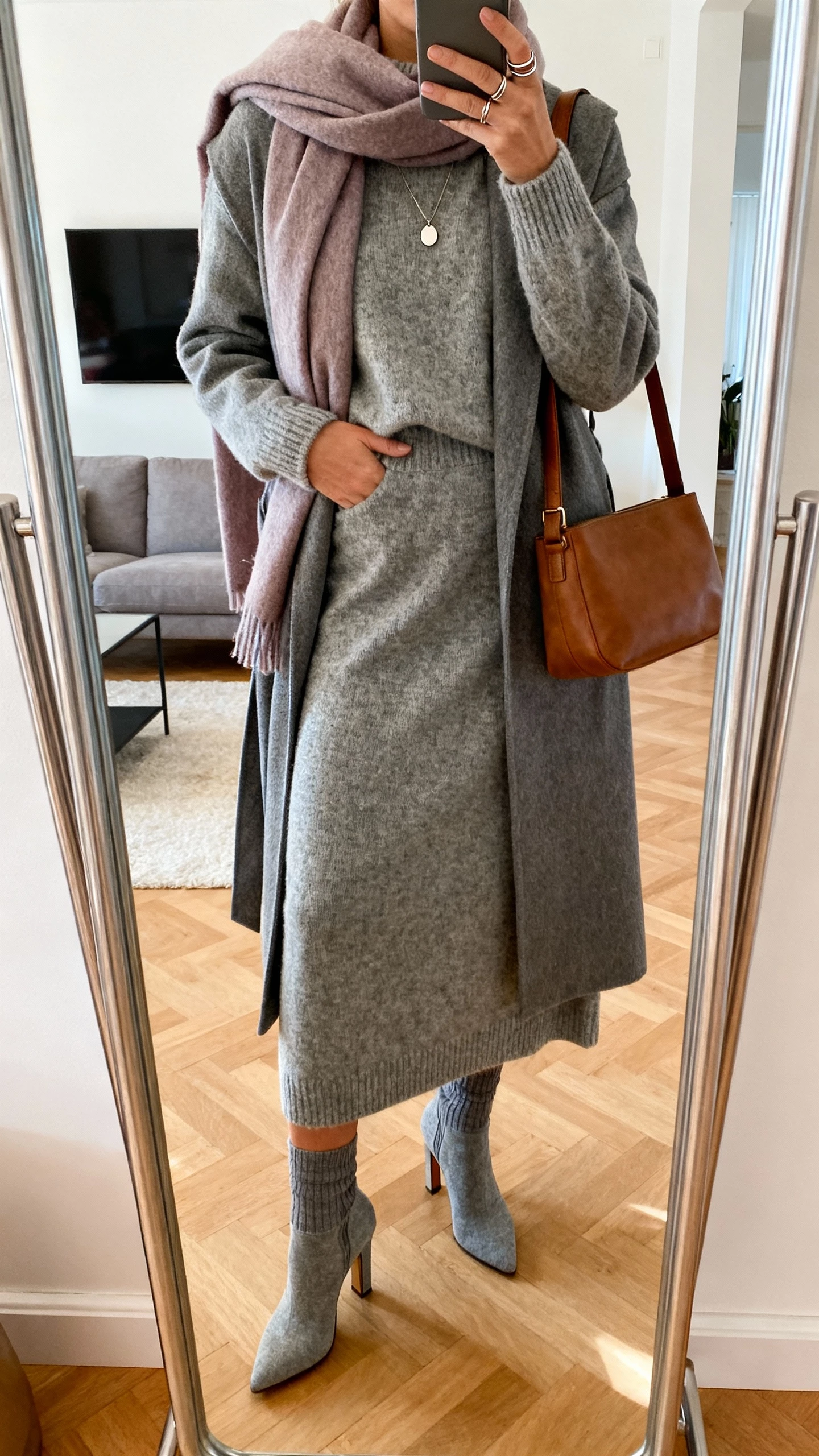 iPhone mirror selfie of a woman in a heather gray knit sweater and matching midi skirt set, heeled sock boots, longline wrap coat, soft scarf in a complementary tone, leather shoulder bag, simple pendant necklace and thin stacking rings, face not visible, living room full-length mirror, warm indoor lighting, natural iPhone photo quality.