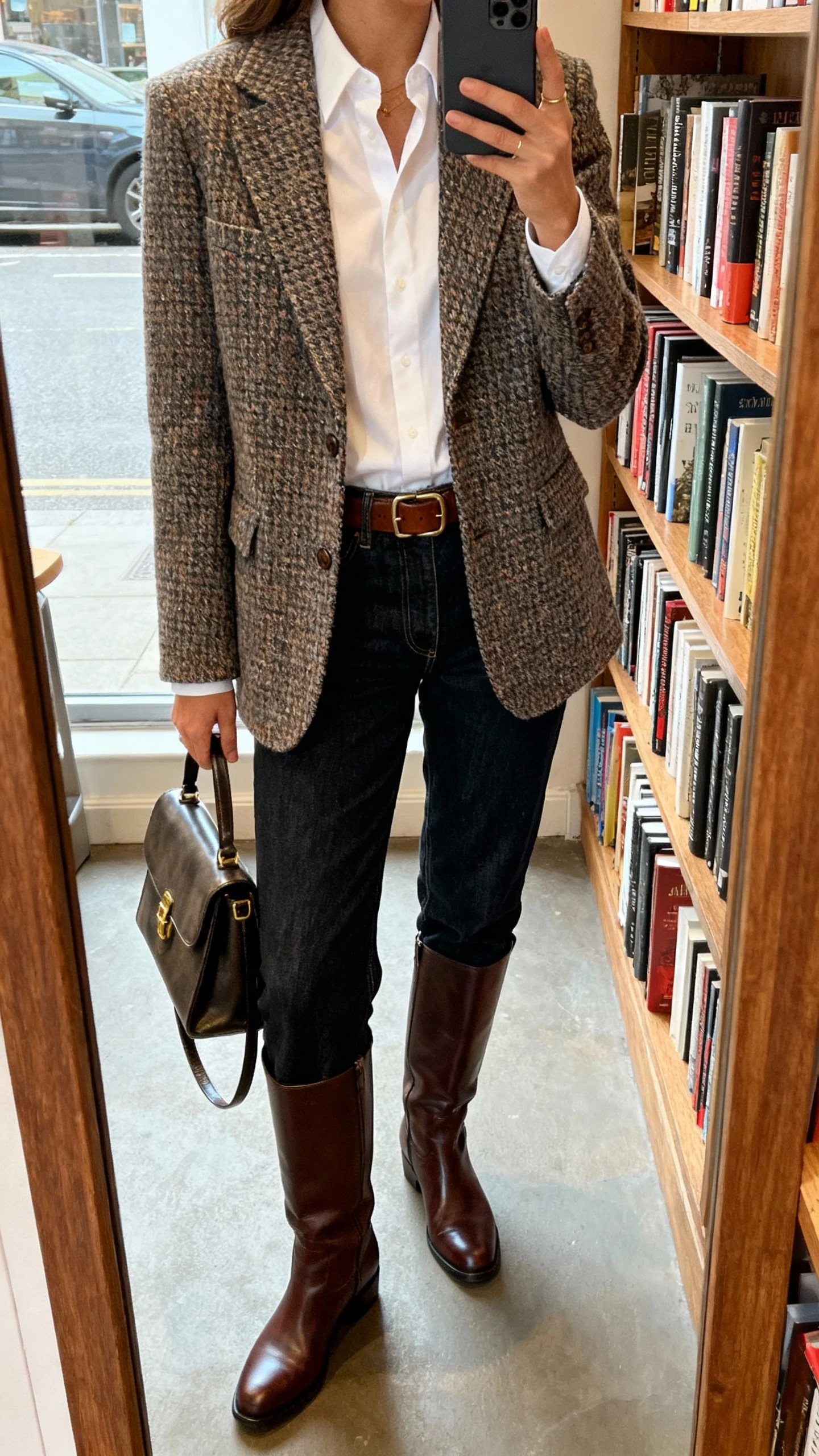 iPhone mirror selfie of a woman in a heritage mix: tweed blazer over a crisp white shirt, straight-leg dark denim, leather belt, knee-high leather boots, and a structured satchel, face not visible, bookstore mirror corner, natural daylight.