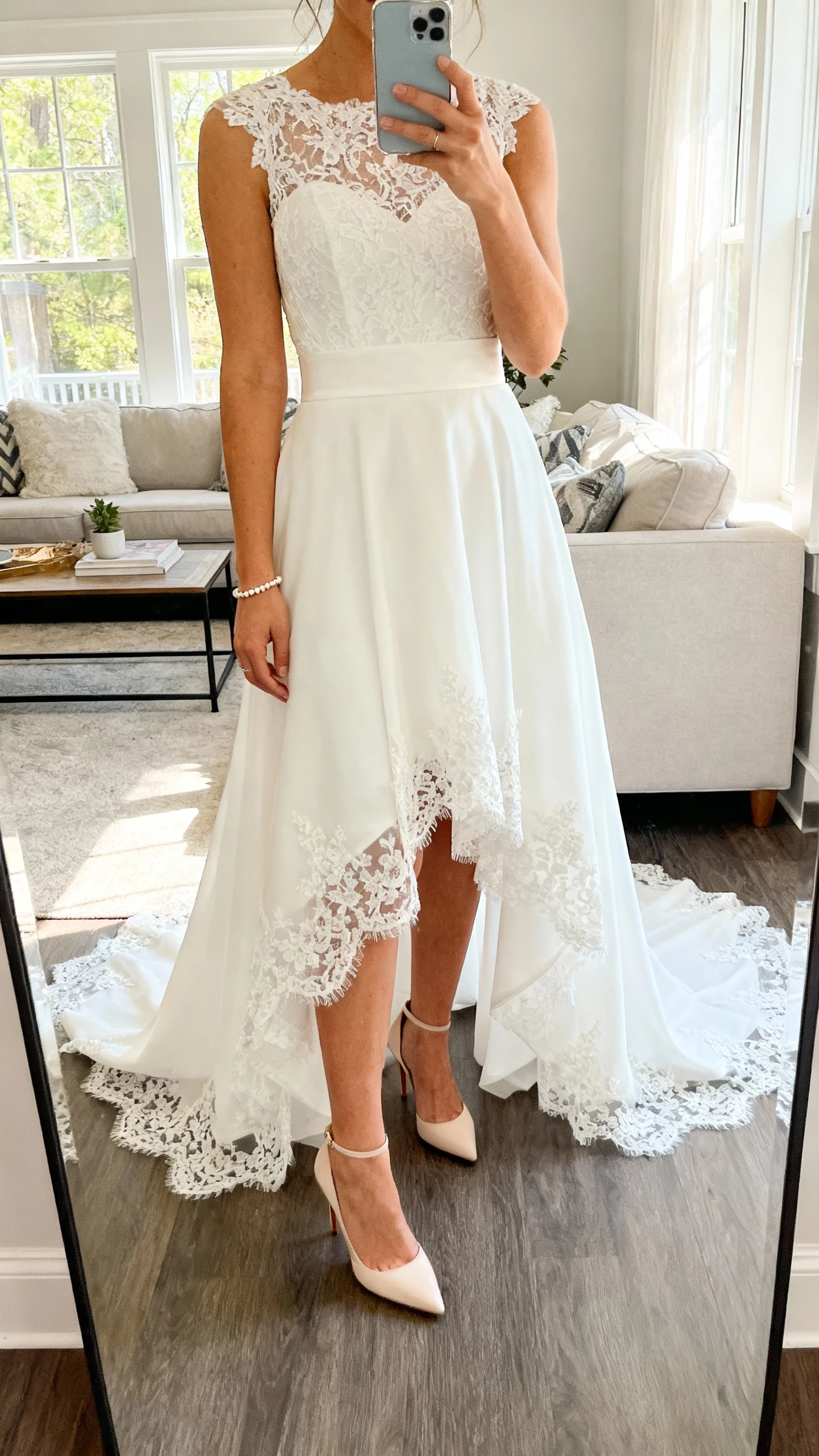 iPhone mirror selfie of a woman in a high-low lace hem wedding dress showing elegant heels, lace bodice with fitted waist, dainty bracelet, face not visible, living room mirror, morning window light, natural iPhone photo quality.