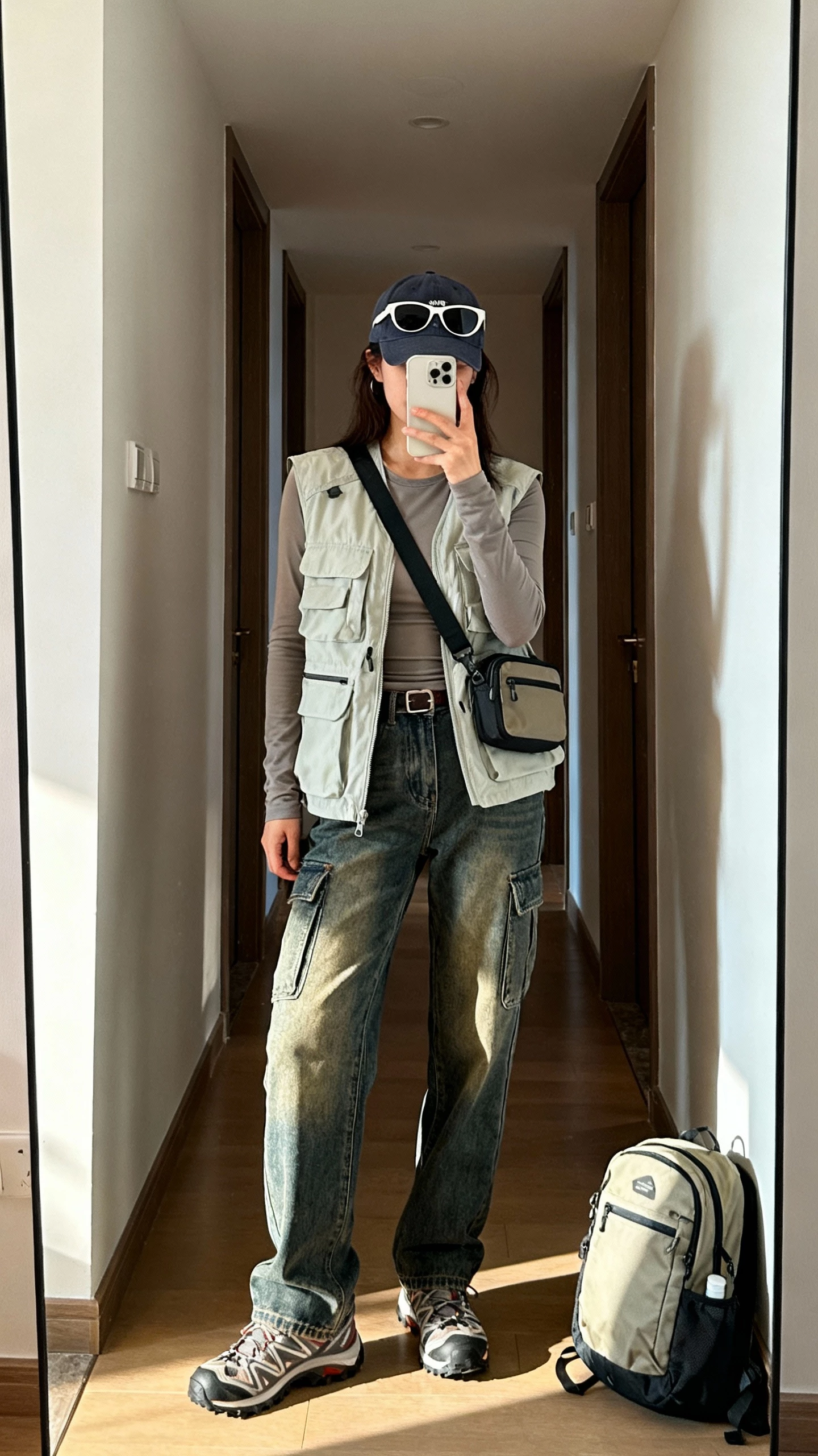 iPhone mirror selfie of a woman in a lightweight utility vest over a fitted long-sleeve tee, classic straight cargo jeans, trail sneakers, hands-free crossbody, compact backpack on floor, baseball cap and subtle aviators, face not visible, apartment hallway mirror, morning light, iPhone photo quality.