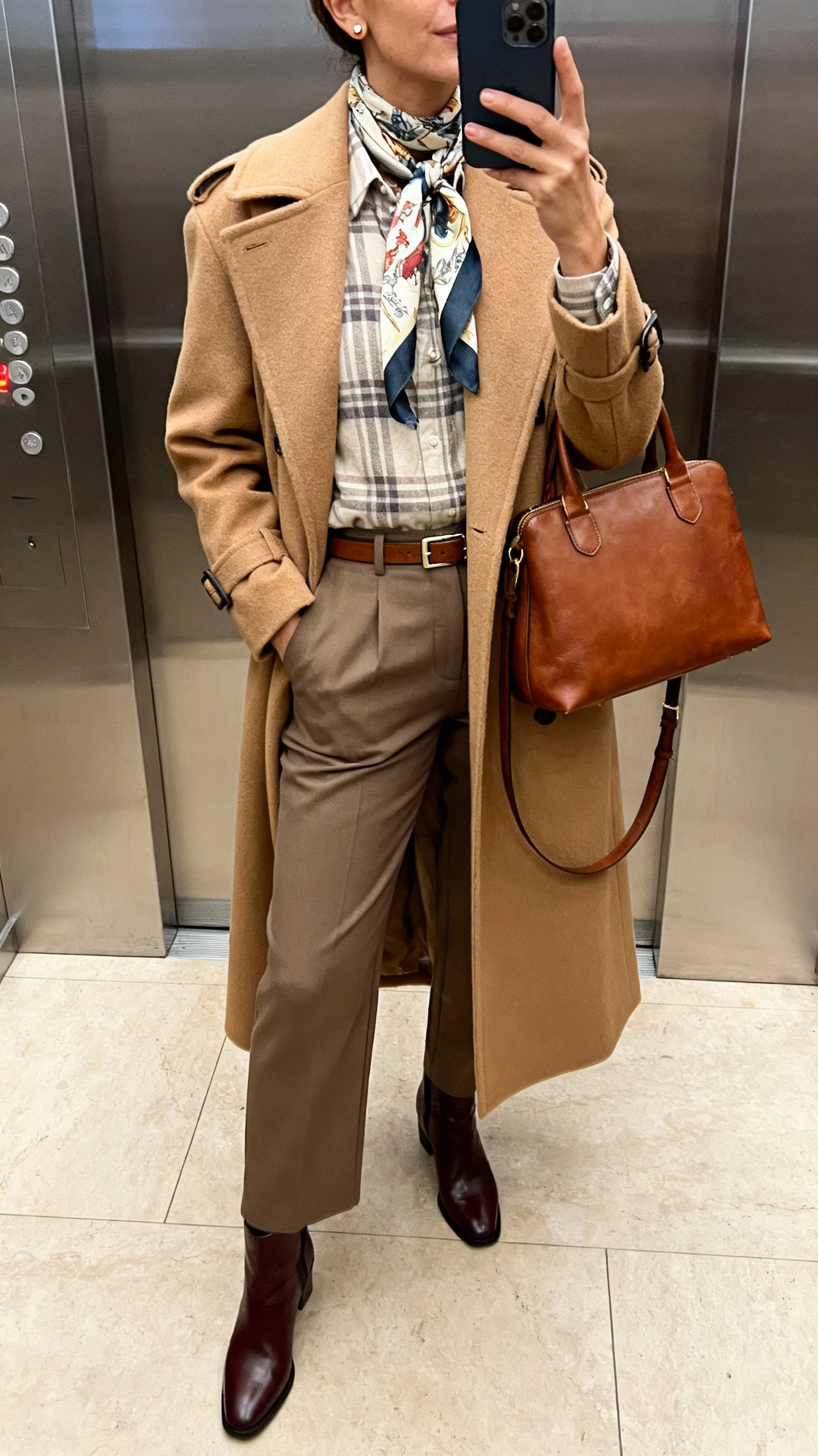 iPhone mirror selfie of a woman in a long camel wool trench, soft plaid flannel shirt, tailored trousers, polished ankle boots, printed silk scarf, leather satchel, minimal stud earrings and a slim belt; elevator mirror, face not visible, indoor lighting, natural iPhone photo quality.