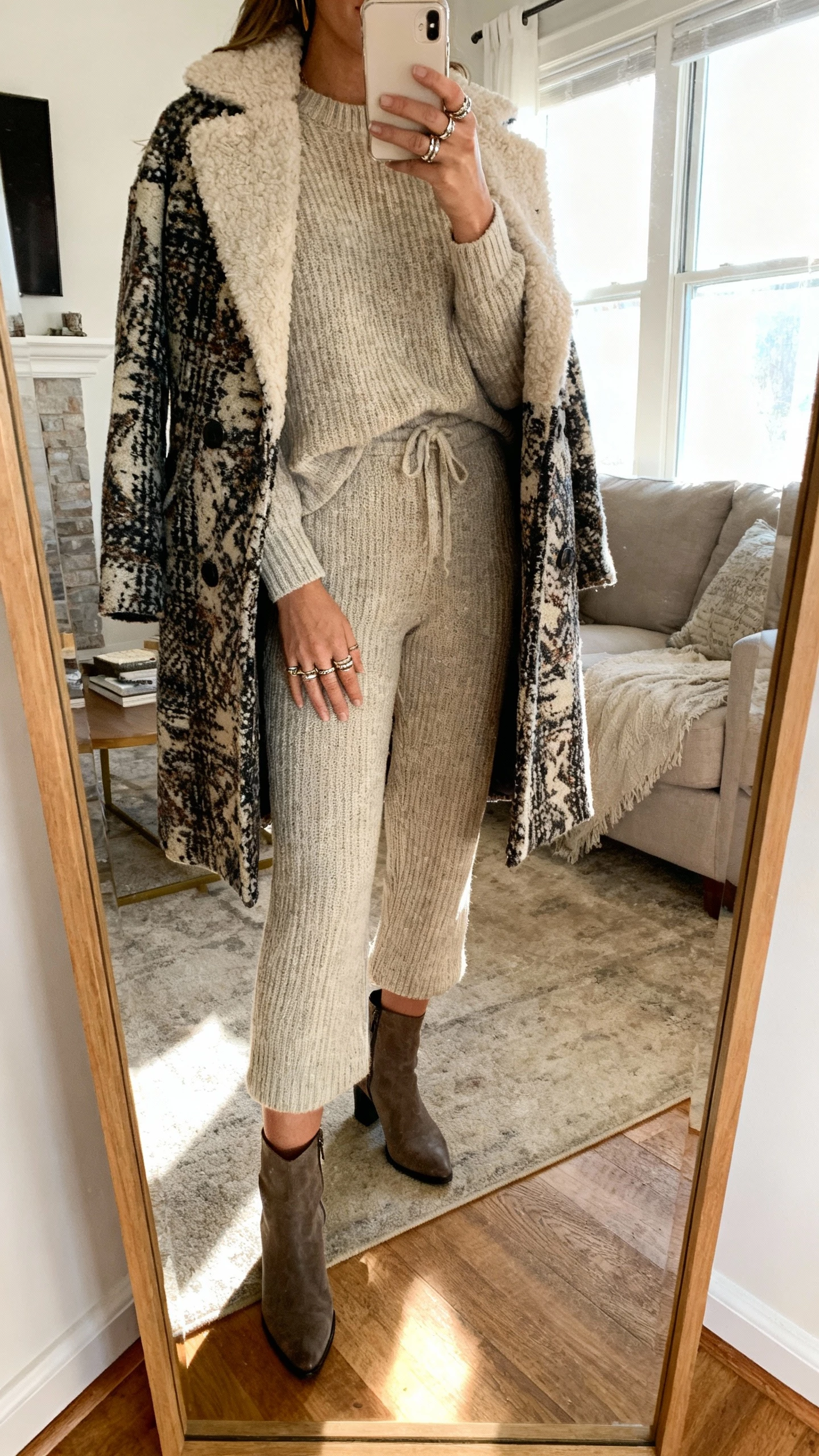 iPhone mirror selfie of a woman in a matching knit set topped with a statement coat, layered rings and ankle boots, face not visible, living room full-length mirror with bright window light, natural iPhone photo quality.