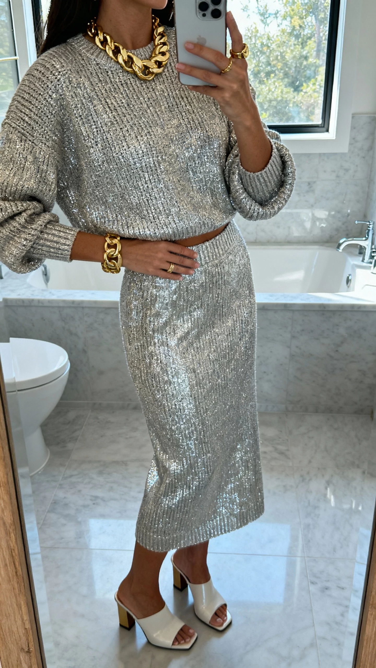 iPhone mirror selfie of a woman in a metallic knit matching set (top and skirt) with chunky gold jewelry and heeled mules, face not visible, bathroom mirror, cool morning light from window, casual iPhone aesthetic.