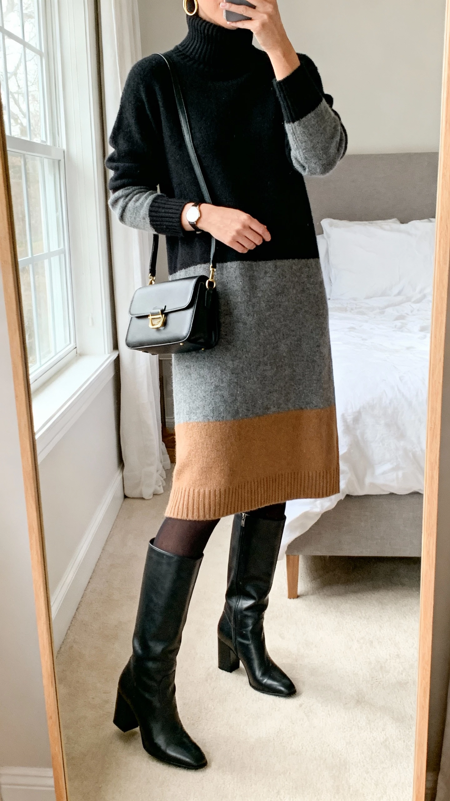 iPhone mirror selfie of a woman in a mid-length cashmere turtleneck sweater dress (black/charcoal/camel) with leather knee-high block-heel boots, structured crossbody or small top-handle tote, minimal gold hoops, slim watch, optional opaque black tights; face not visible, cropped at neck, bedroom mirror, natural window light, natural iPhone photo quality.