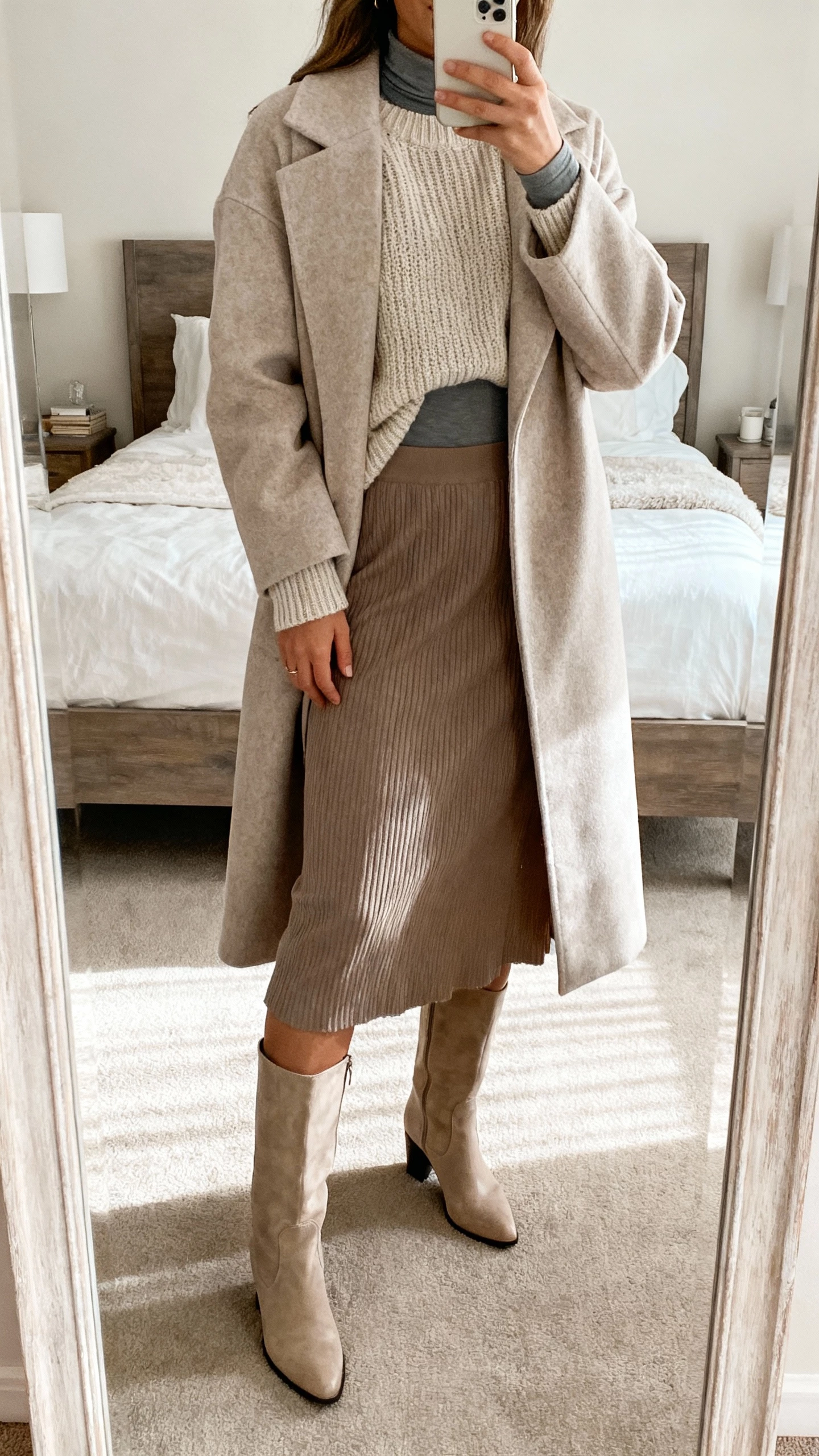 iPhone mirror selfie of a woman in a midi skirt with tall boots and a hidden thermal layer under a knit, long coat over top, face not visible, bedroom mirror, soft daylight, natural iPhone aesthetic.