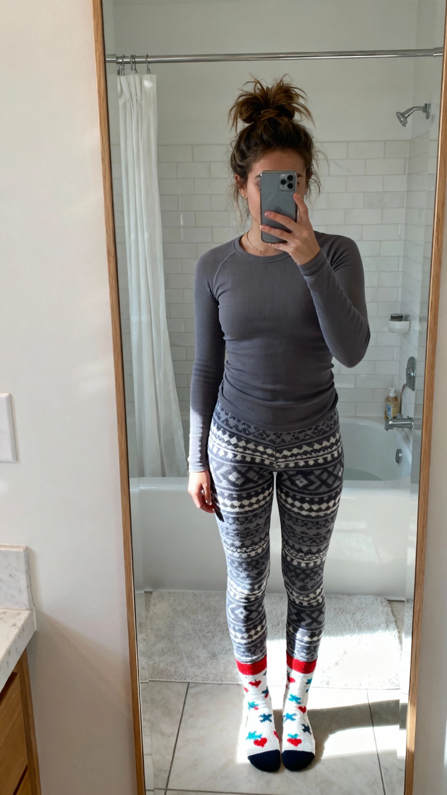 iPhone mirror selfie of a woman in a mix-and-match thermal set (solid thermal top and patterned thermal pants) with playful socks and hair in a messy bun, face not visible, bathroom mirror, bright natural light, natural iPhone photo quality.