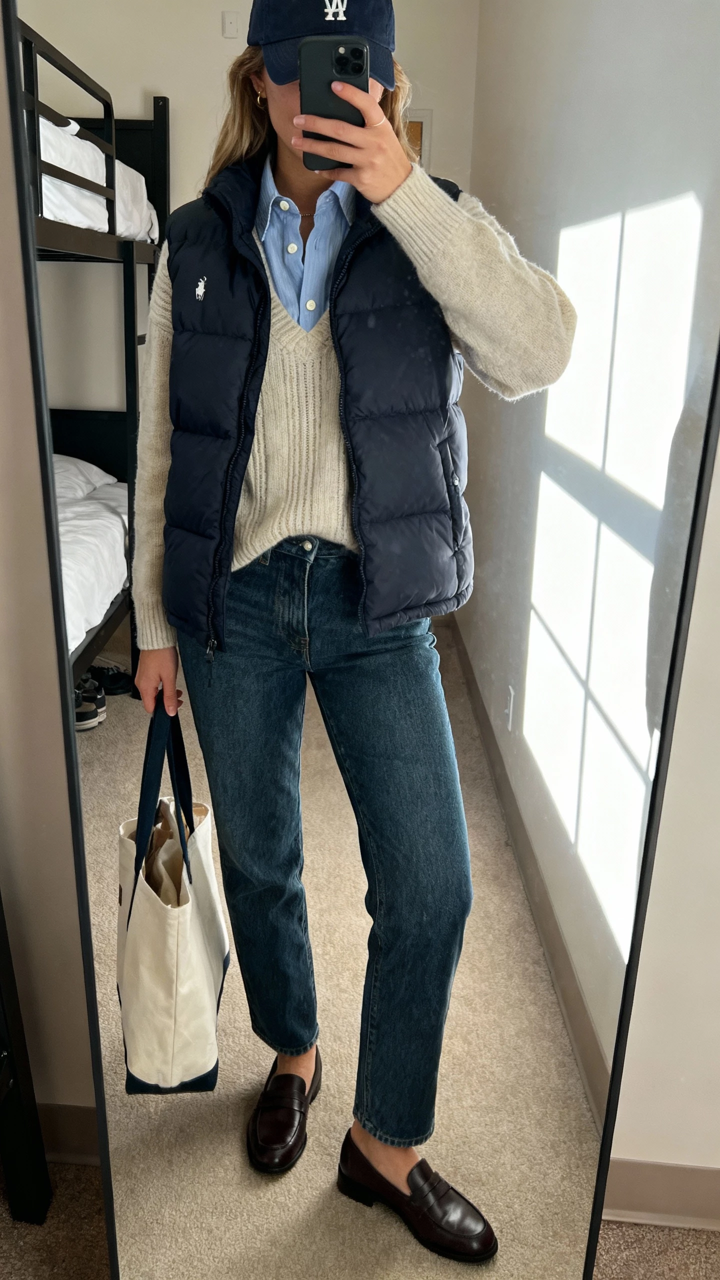 iPhone mirror selfie of a woman in a preppy puffer vest layered over a button-down and fine-knit sweater, straight jeans, loafers, baseball cap, tote, face not visible, dorm-style bedroom mirror with window light, natural iPhone photo quality.