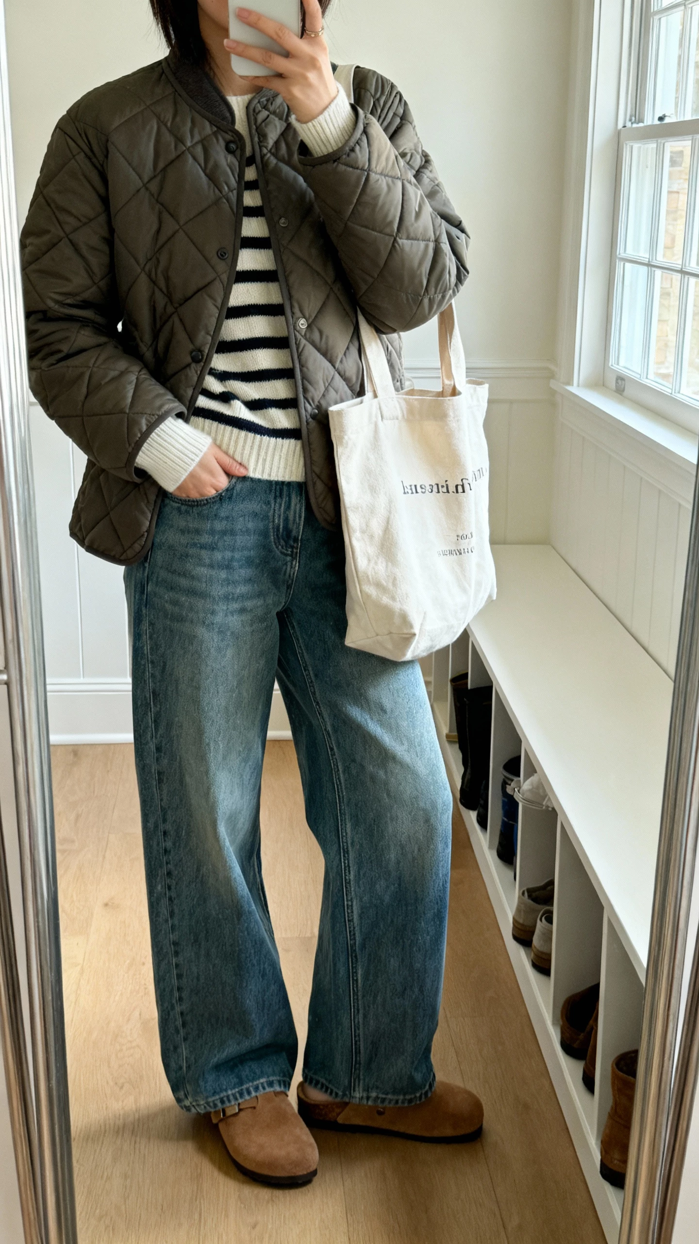 iPhone mirror selfie of a woman in a quilted jacket with wide-leg jeans, striped knit underneath, suede clogs, canvas tote, face not visible, mudroom mirror, bright daytime window light, natural iPhone photo quality