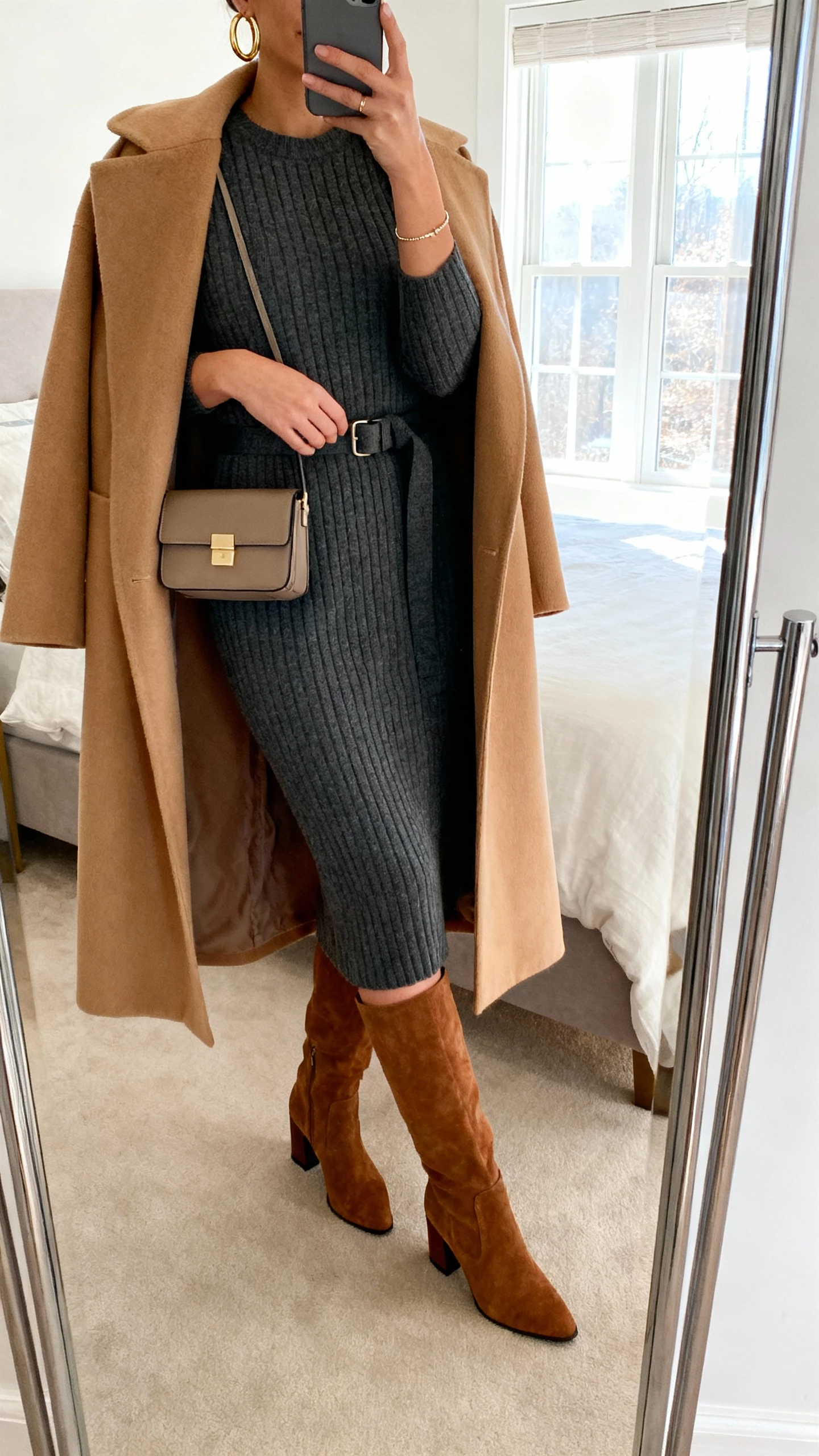 iPhone mirror selfie of a woman in a ribbed knit midi dress (charcoal) with knee-high suede block-heel boots, optional belt, long camel wool coat draped, structured mini bag, gold hoop earrings and delicate bracelet, face not visible, cropped at neck, bedroom mirror with natural window light, natural iPhone photo quality.
