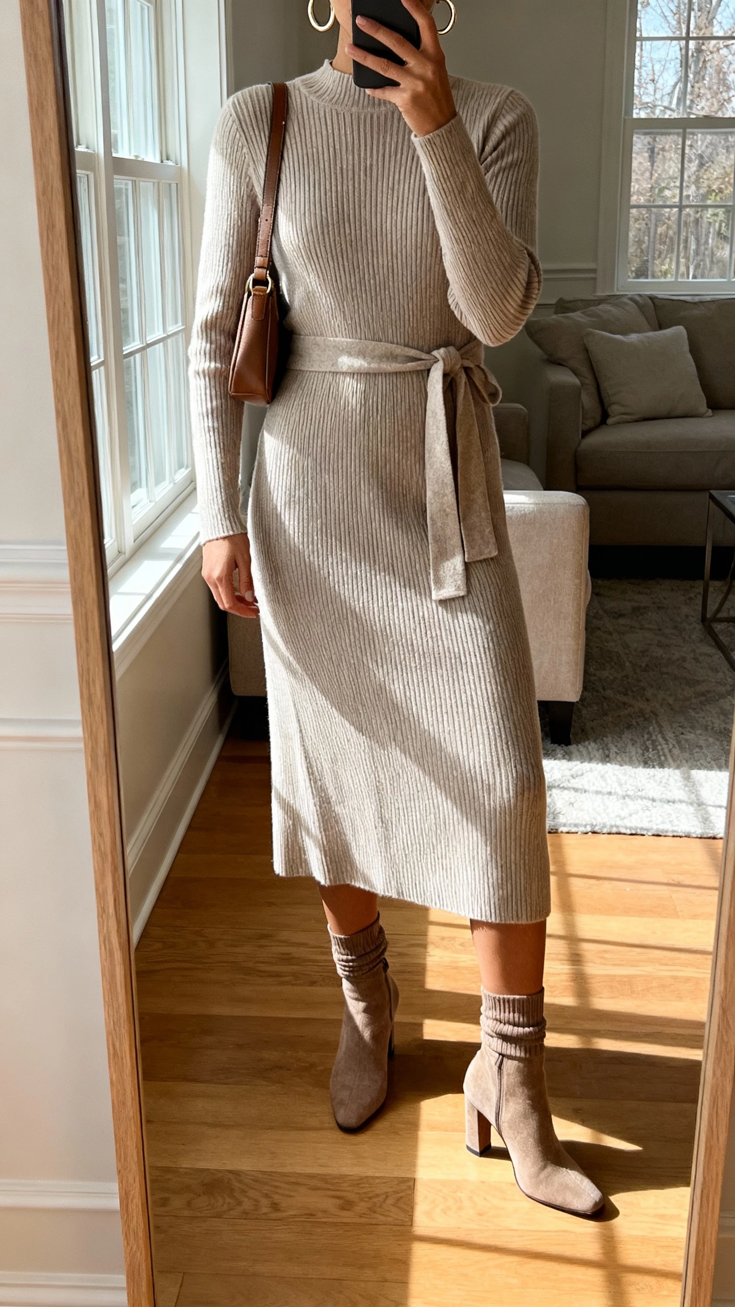 iPhone mirror selfie of a woman in a ribbed knit midi dress with a waist tie, heeled sock boots, hoop earrings, crossbody bag, face not visible, cropped at shoulders, living room mirror with afternoon window light, natural iPhone aesthetic.