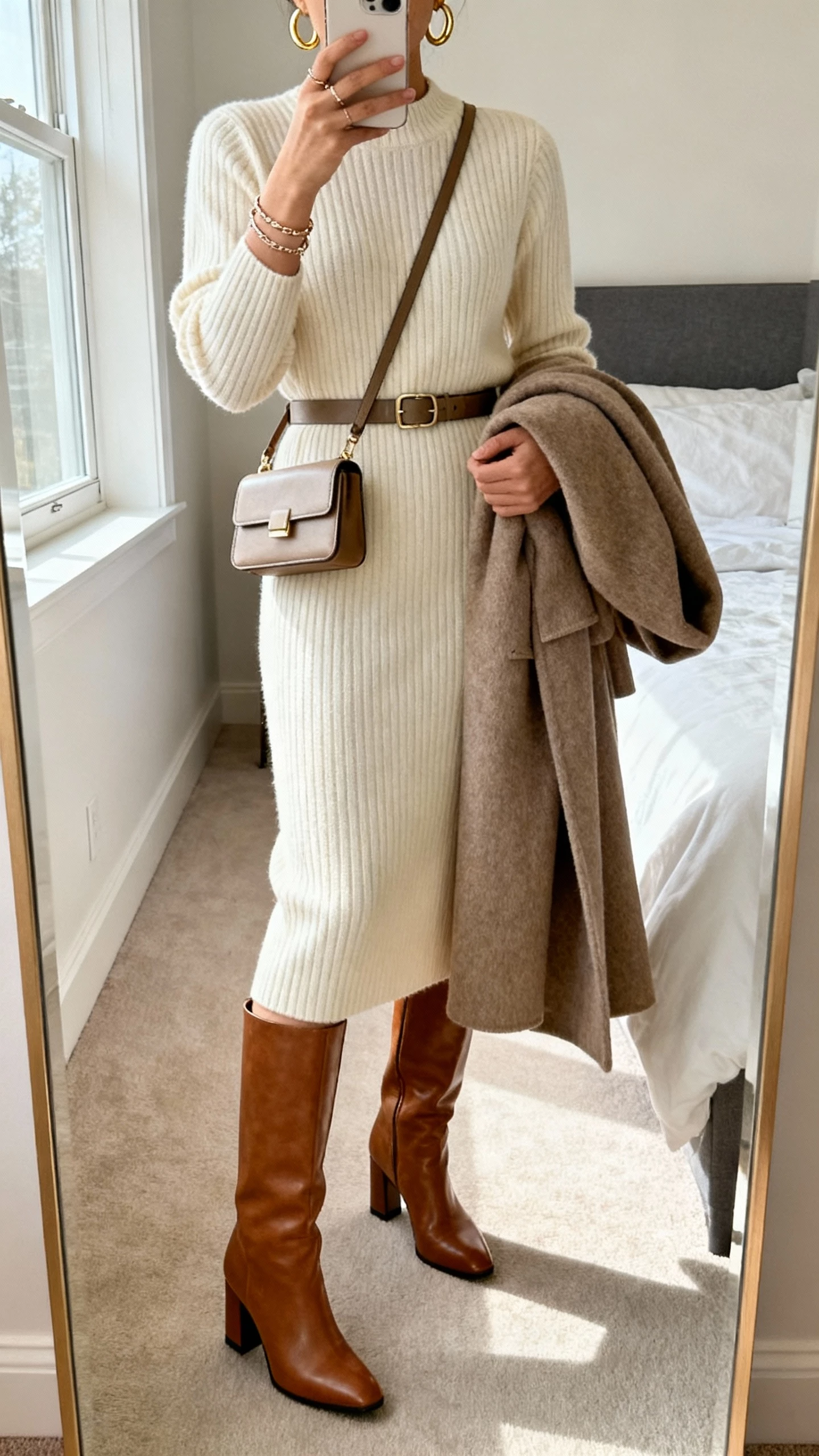 iPhone mirror selfie of a woman in a ribbed midi sweater dress (cream), knee-high leather block-heel boots, sleek waist belt, gold hoop earrings with a delicate bracelet stack, structured mini shoulder bag, and a long wool coat draped over arm; bedroom full-length mirror, face not visible, natural window light, natural iPhone photo quality.