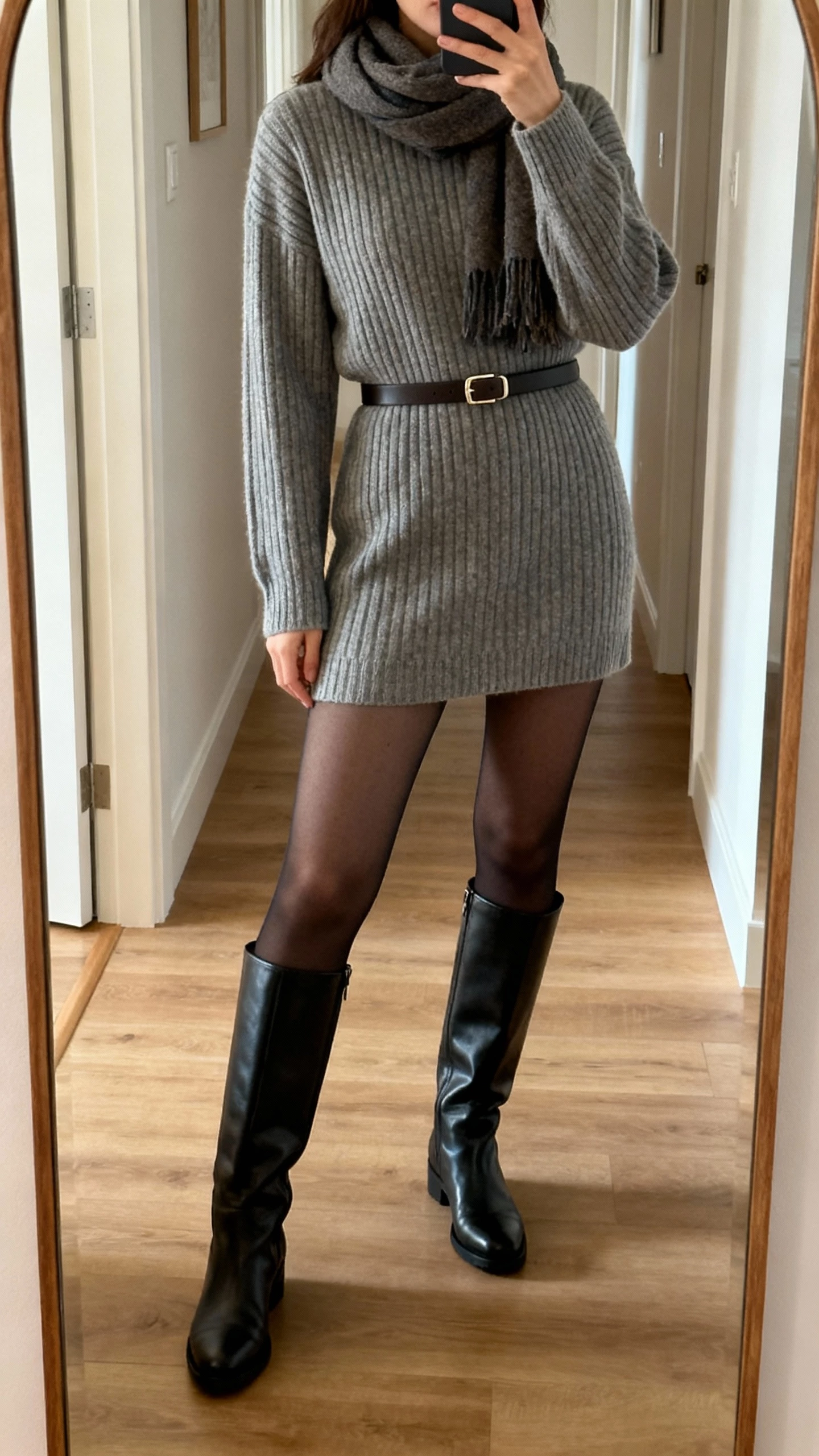 iPhone mirror selfie of a woman in a ribbed winter sweater dress with opaque tights, knee-high boots, a slim belt, and a scarf, face not visible, cropped at shoulders, hallway mirror, soft indoor lighting, natural iPhone quality.