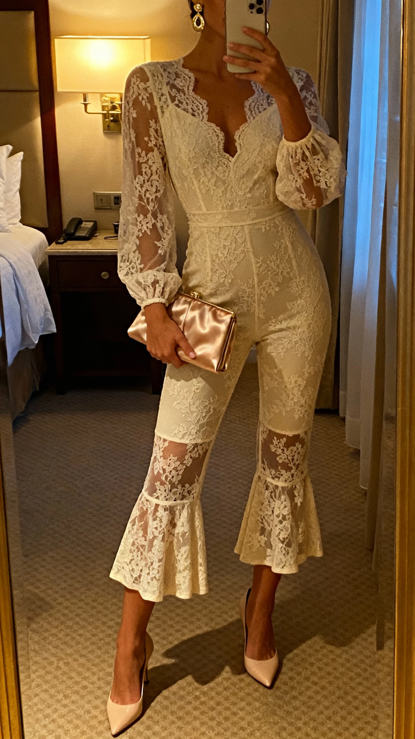 iPhone mirror selfie of a woman in a romantic lace-overlay jumpsuit with cropped kick-flare legs, kitten heels, vintage-inspired earrings, satin clutch, face not visible, boutique hotel room mirror, warm evening lamp light, natural iPhone photo quality