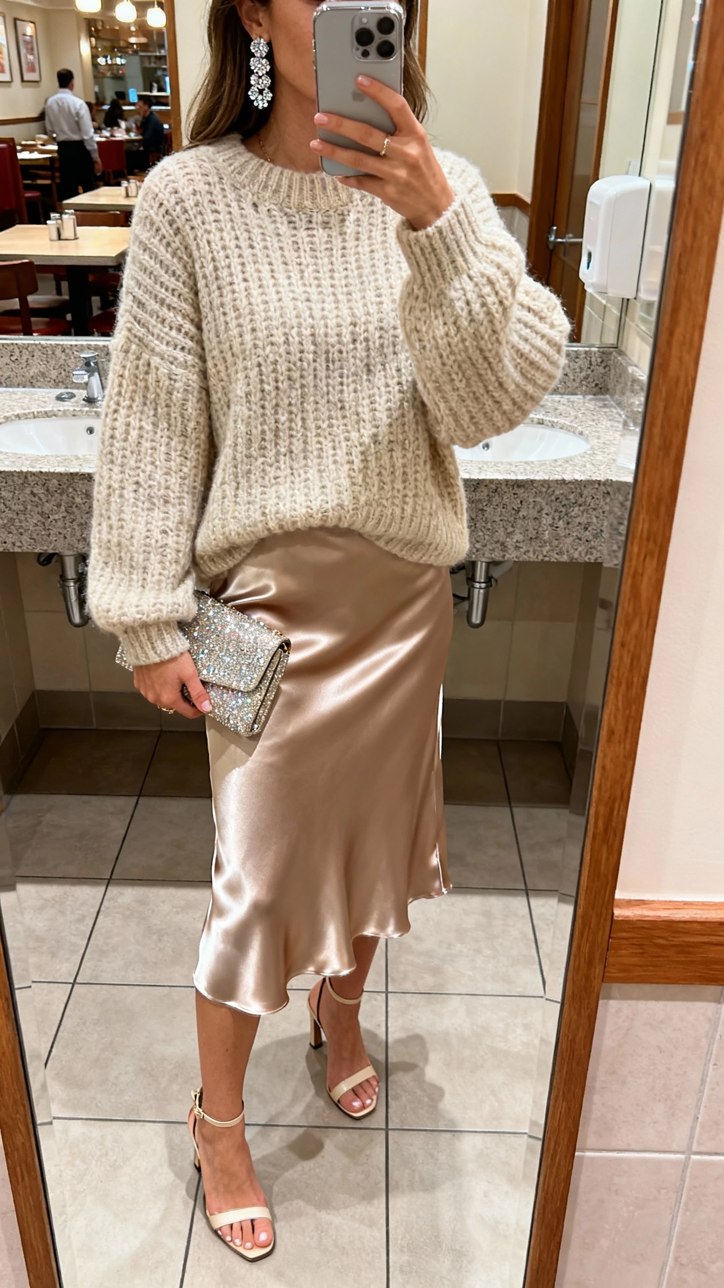 iPhone mirror selfie of a woman in a satin skirt with a chunky knit sweater, sparkly earrings, a glitzy clutch, and heeled sandals, face not visible, bathroom mirror at a restaurant, soft indoor lighting, natural iPhone photo quality